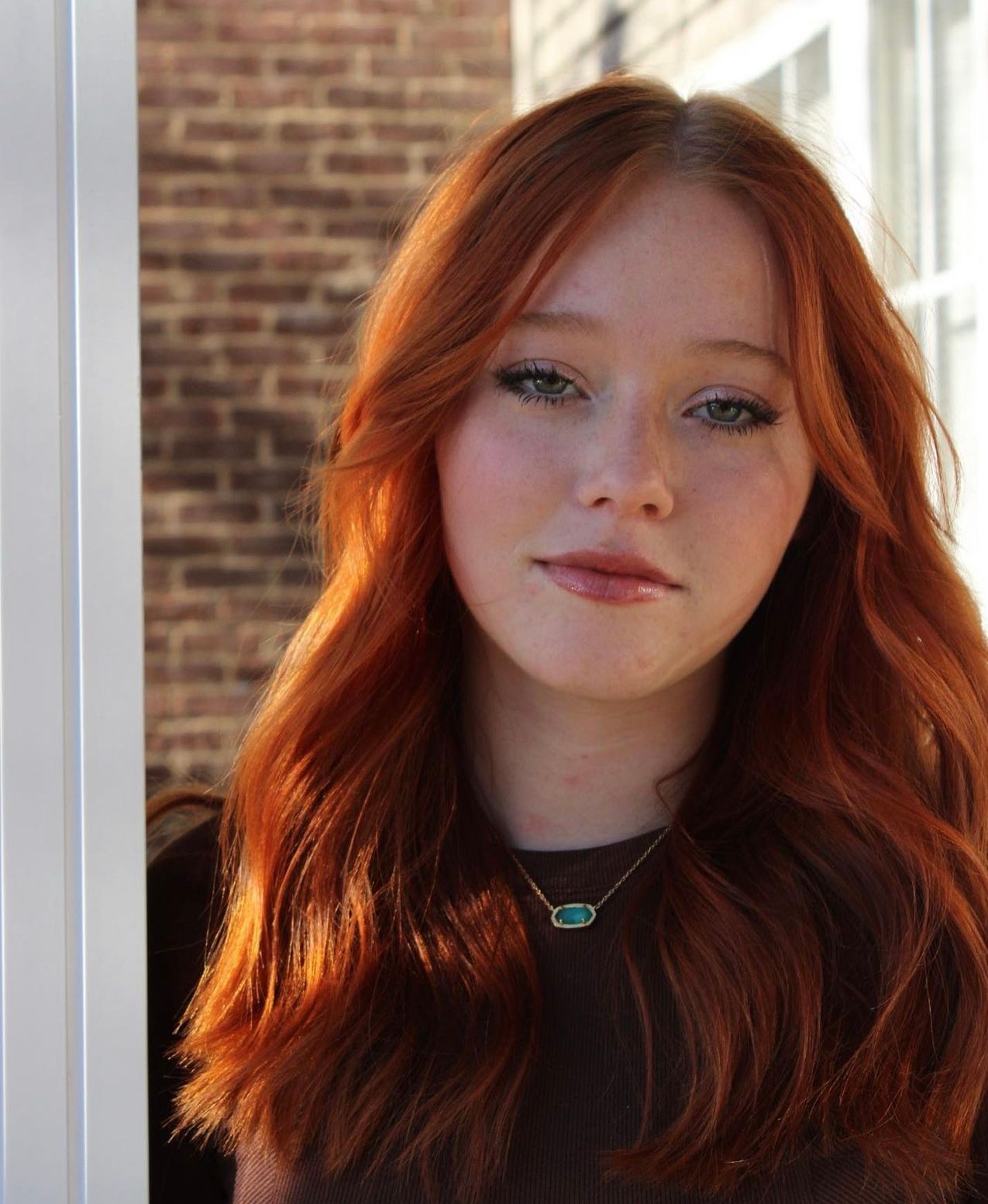 Young woman with long vibrant red hair and freckles posing near a brick wall in natural light.