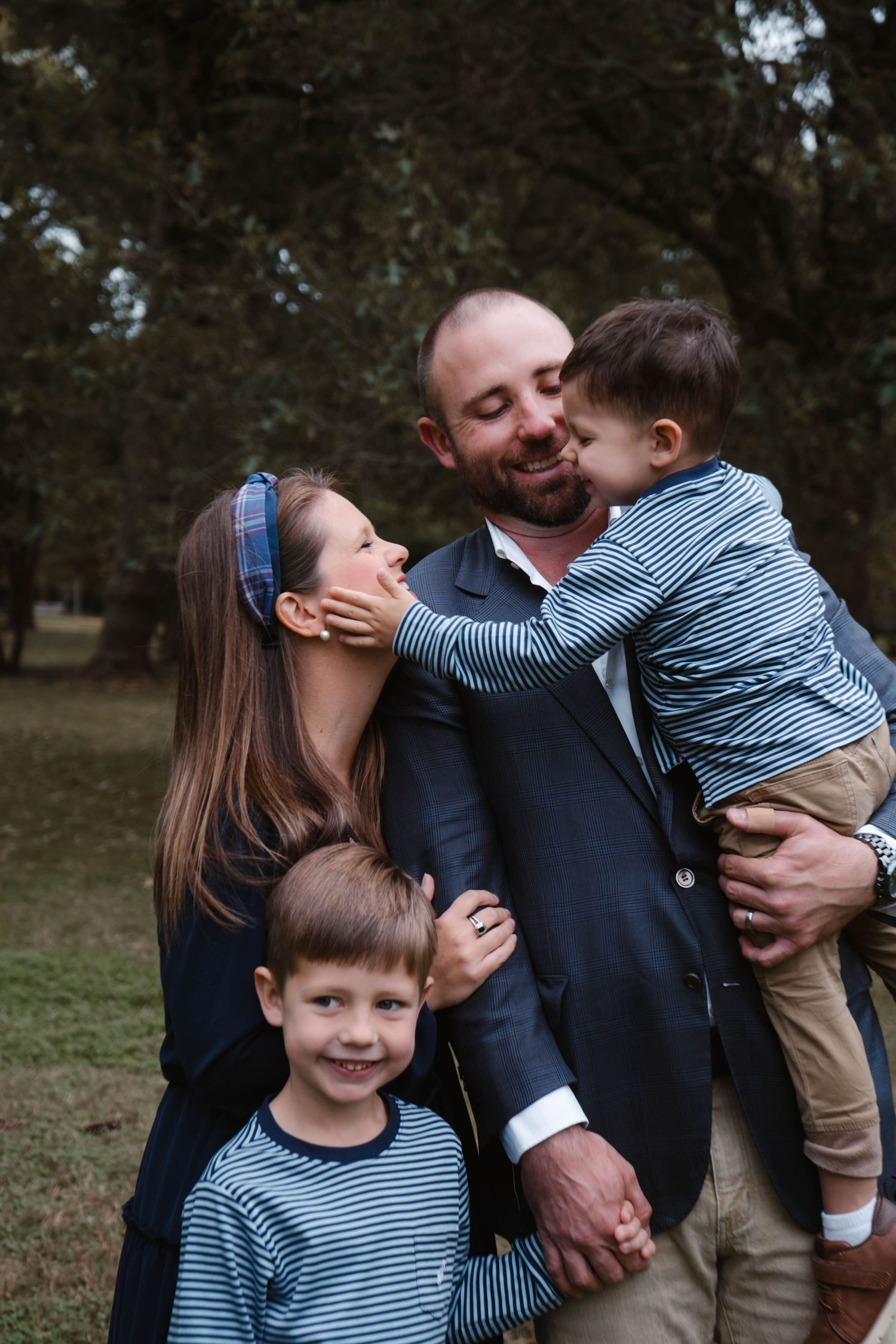 Cheerful family portrait outdoors as toddler kisses fathers cheek while older brother smiles and mother laughs in park.