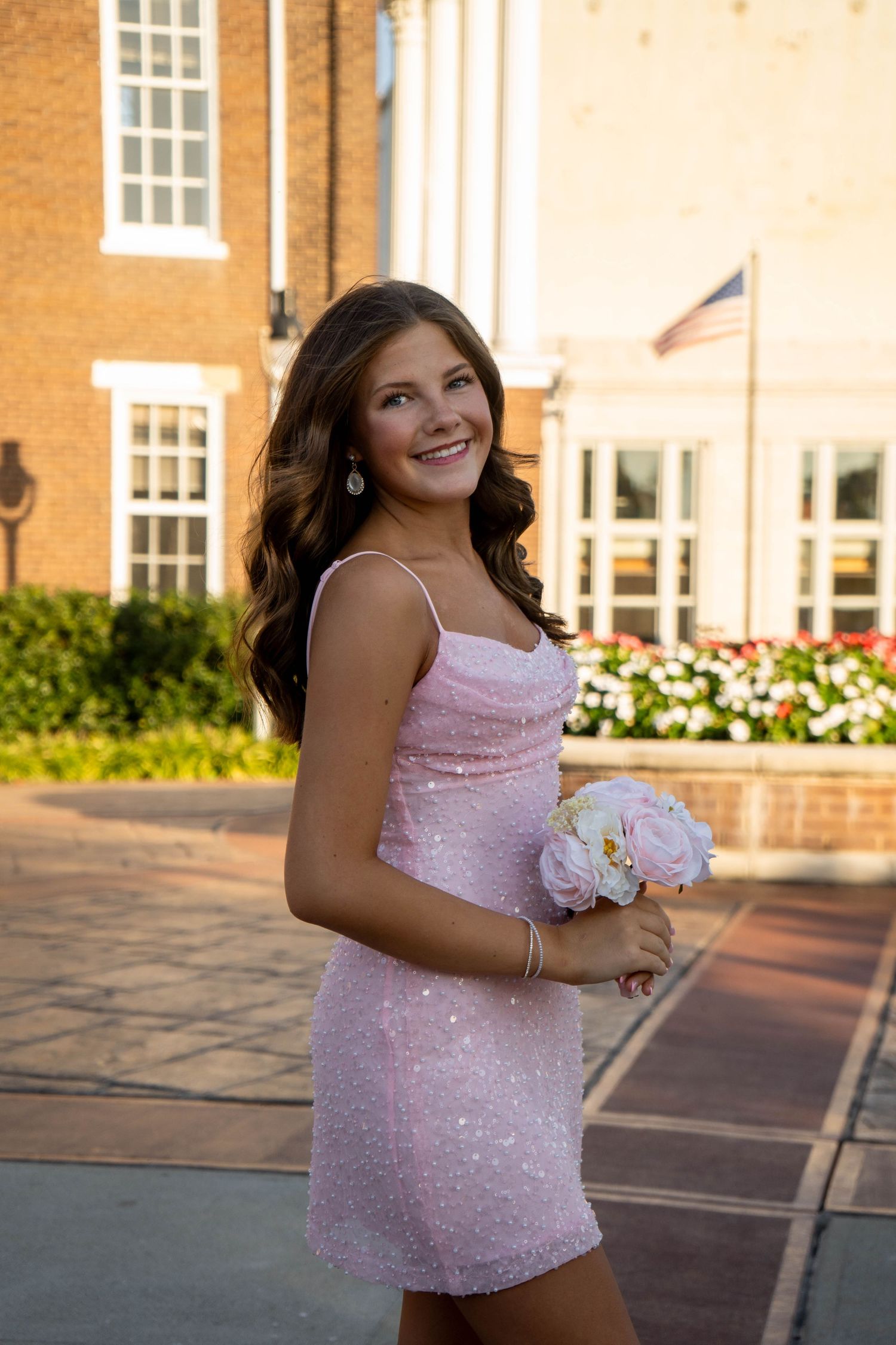 A teenage girl in a sparkly pink mini dress holds a white bouquet while smiling outdoors.