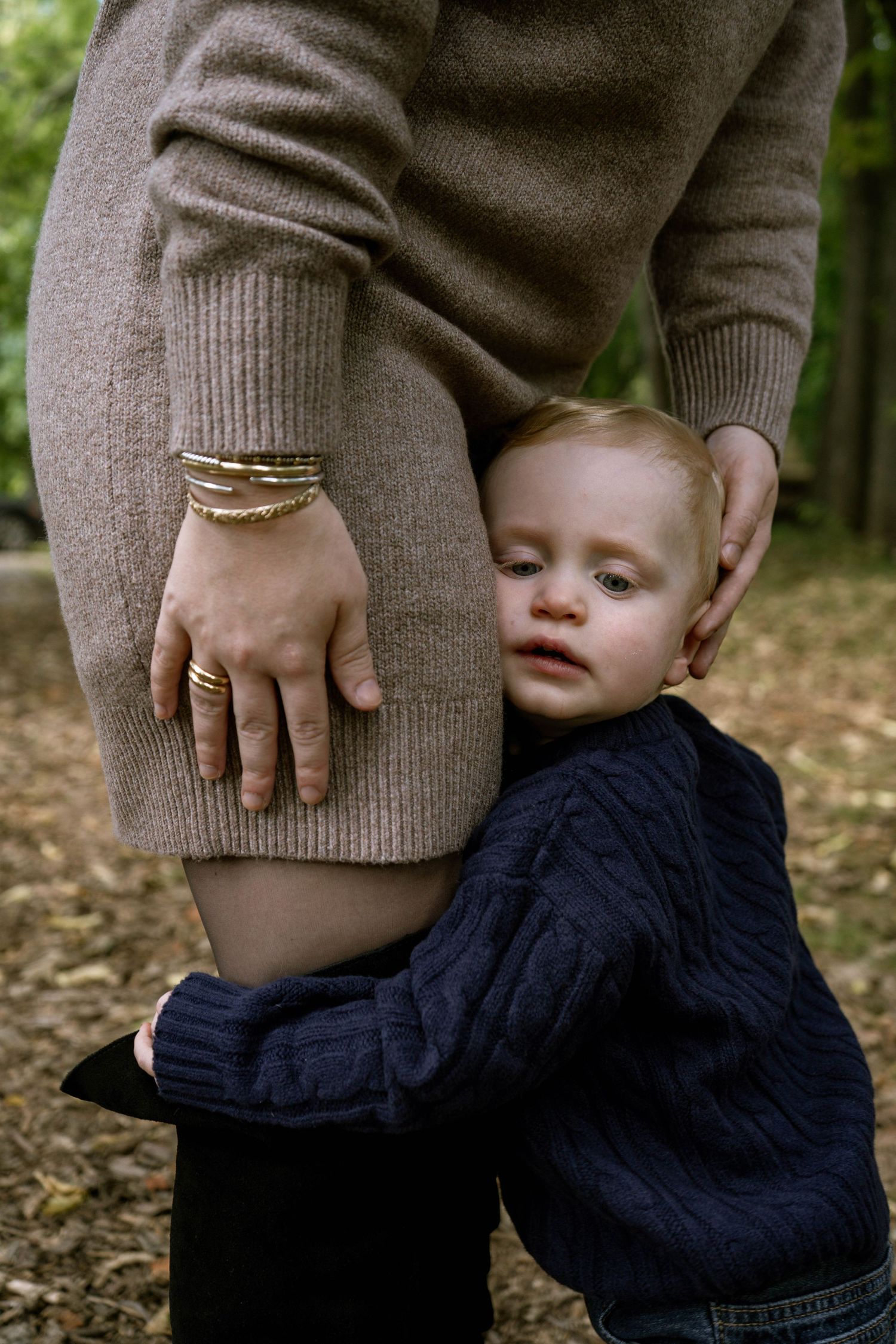 Toddler in navy sweater hugging mother's legs while she gently cradles his head in an outdoor park.