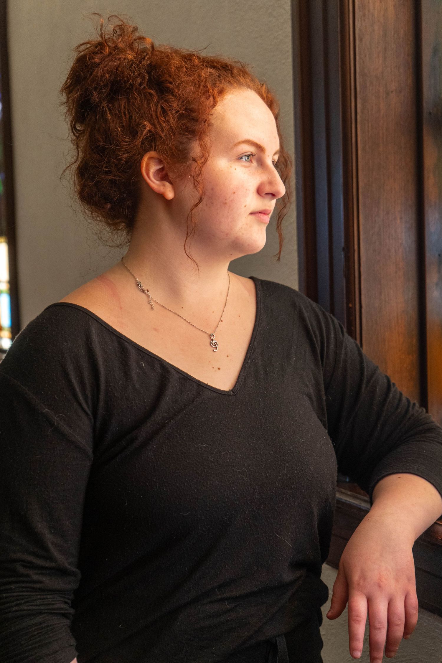 Young woman with red curly hair in black top gazes thoughtfully out window wearing delicate necklace.