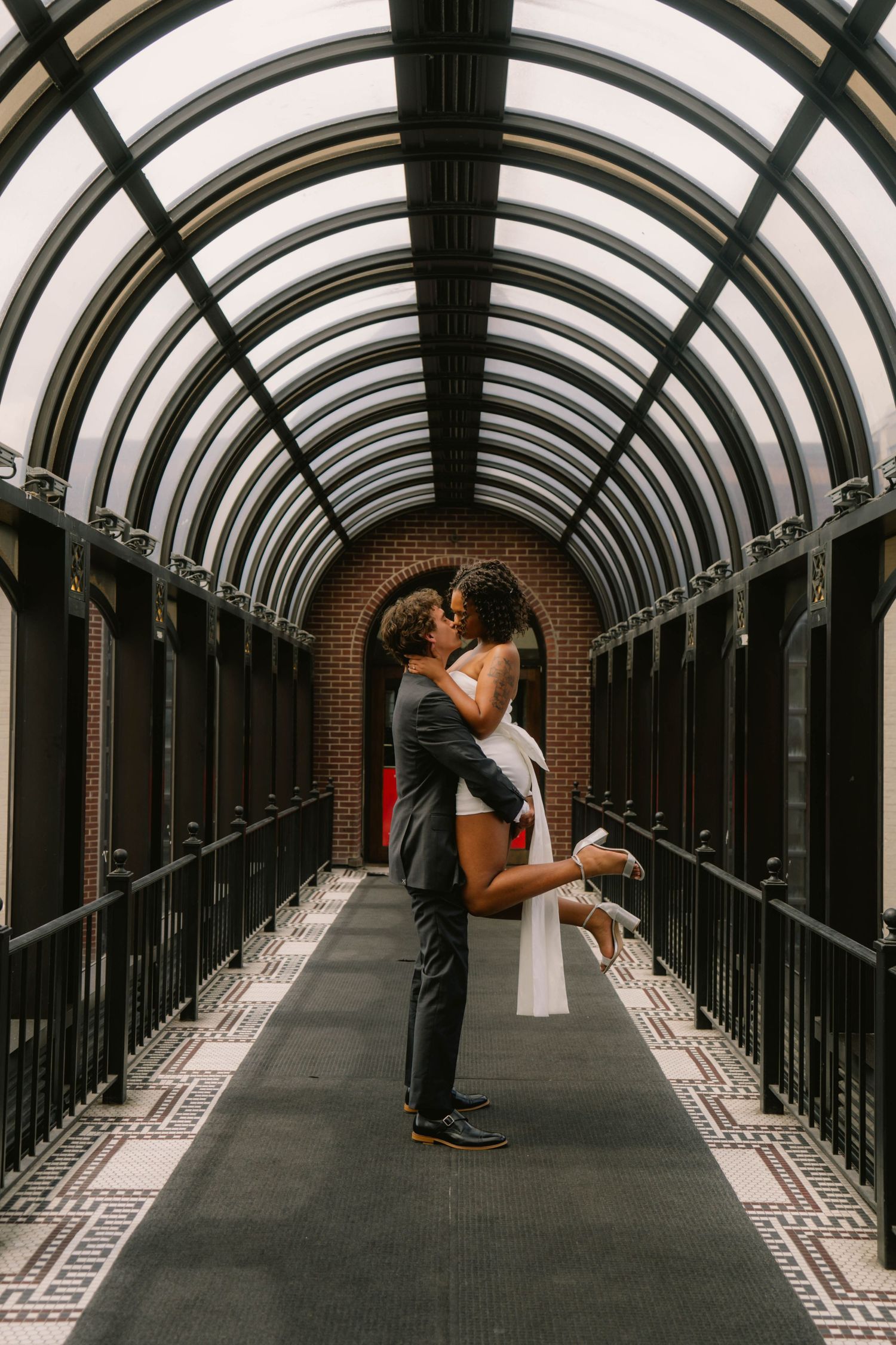Groom lifting bride in white mini dress sharing romantic kiss in elegant glass-roofed tunnel corridor.