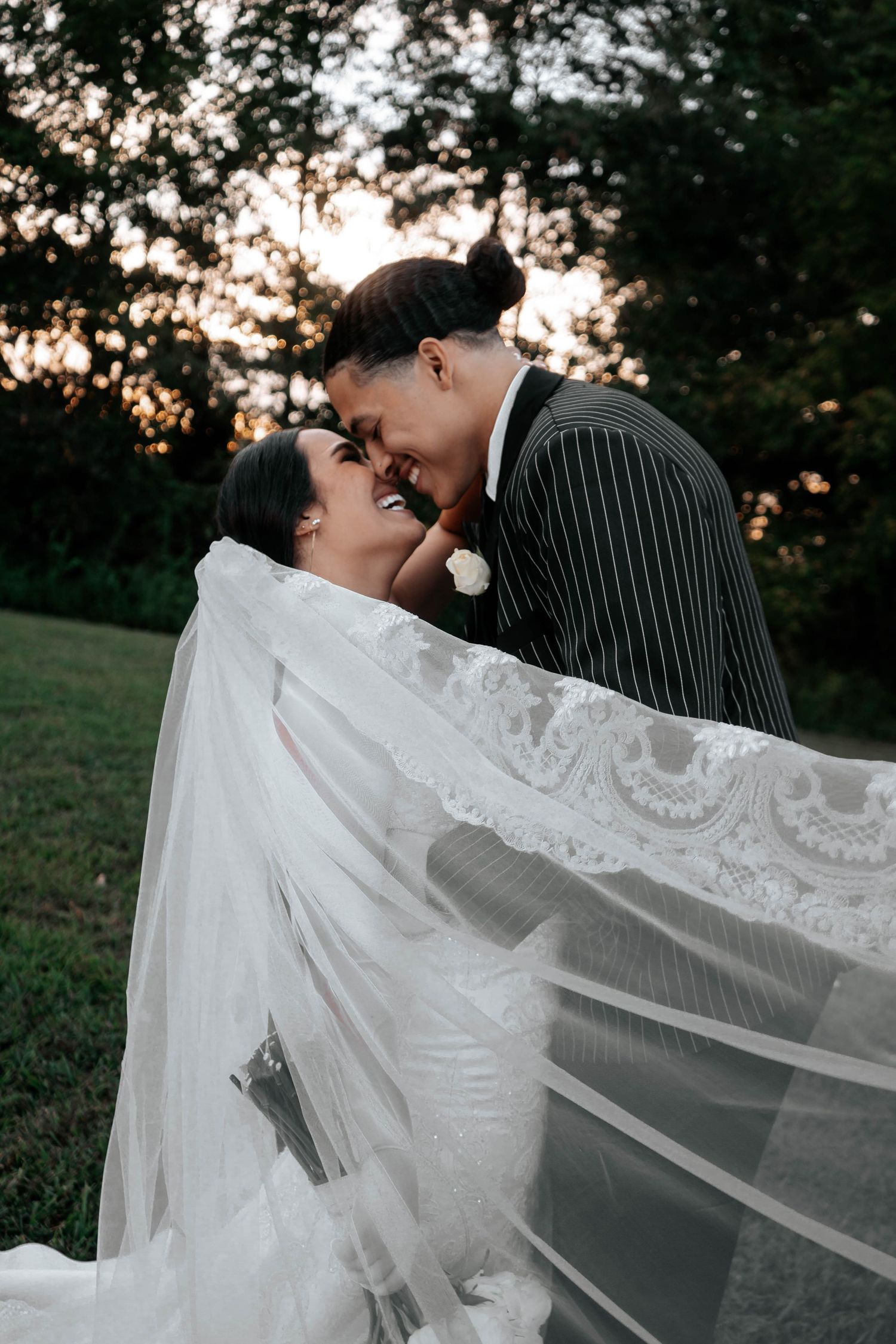 Bride and groom share an intimate nose-to-nose moment outdoors at sunset, with flowing lace wedding veil draped elegantly.