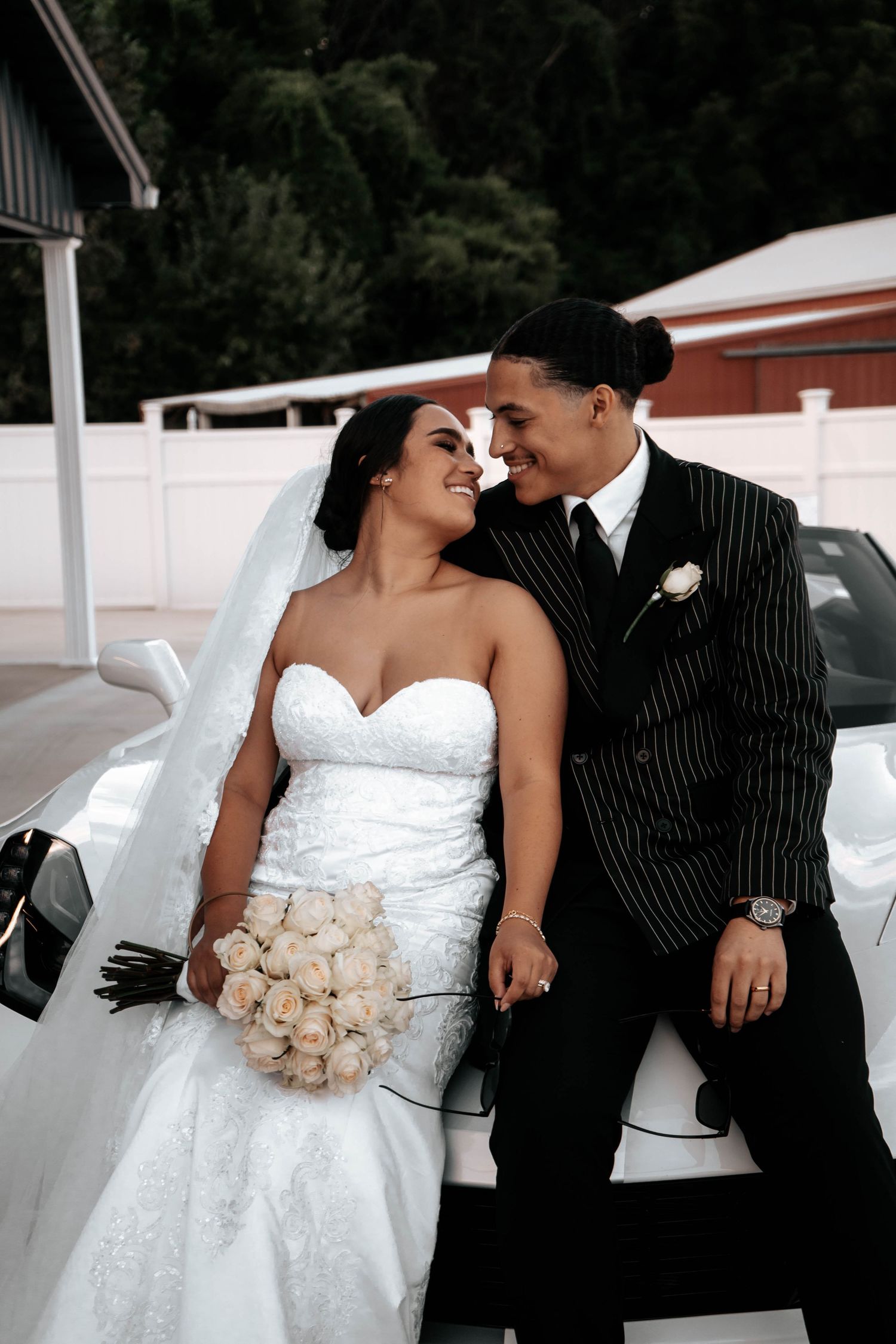 Bride and groom share a romantic moment leaning against a white car, bride holding white rose bouquet.