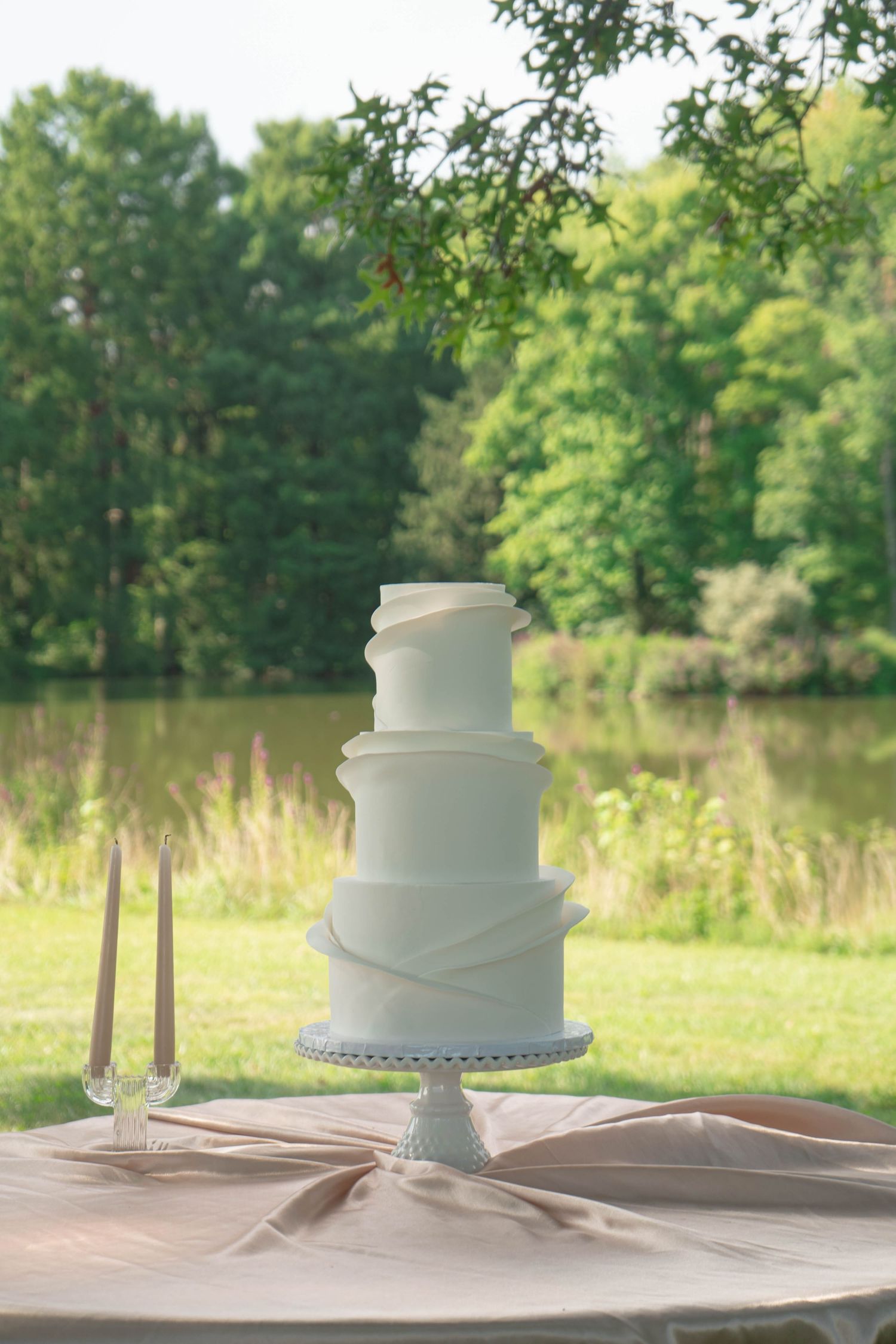 Three-tier white wedding cake with draped fondant detail displayed outdoors by a serene lake.