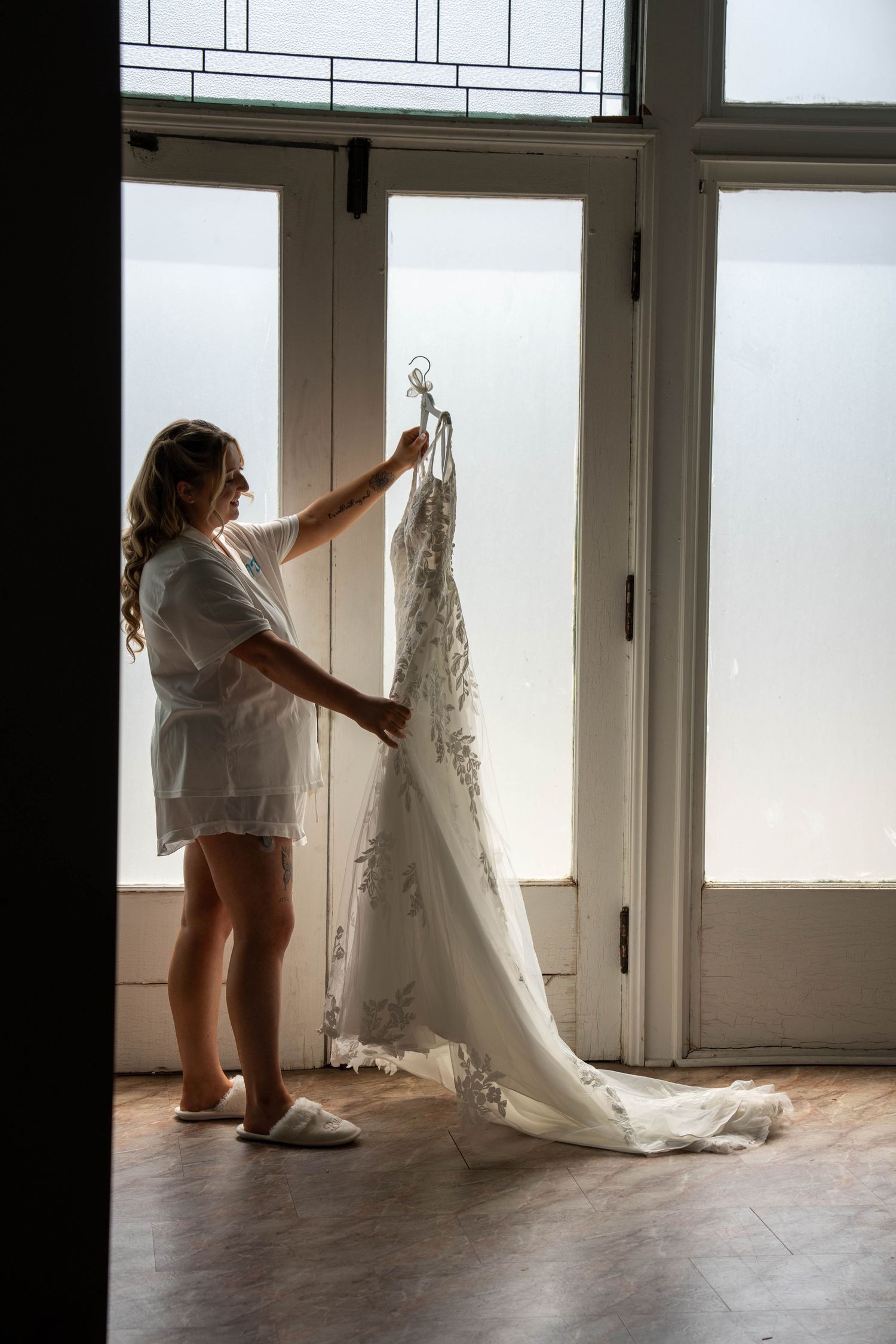 Bride in white robe holding wedding dress on hanger near large window with natural light.