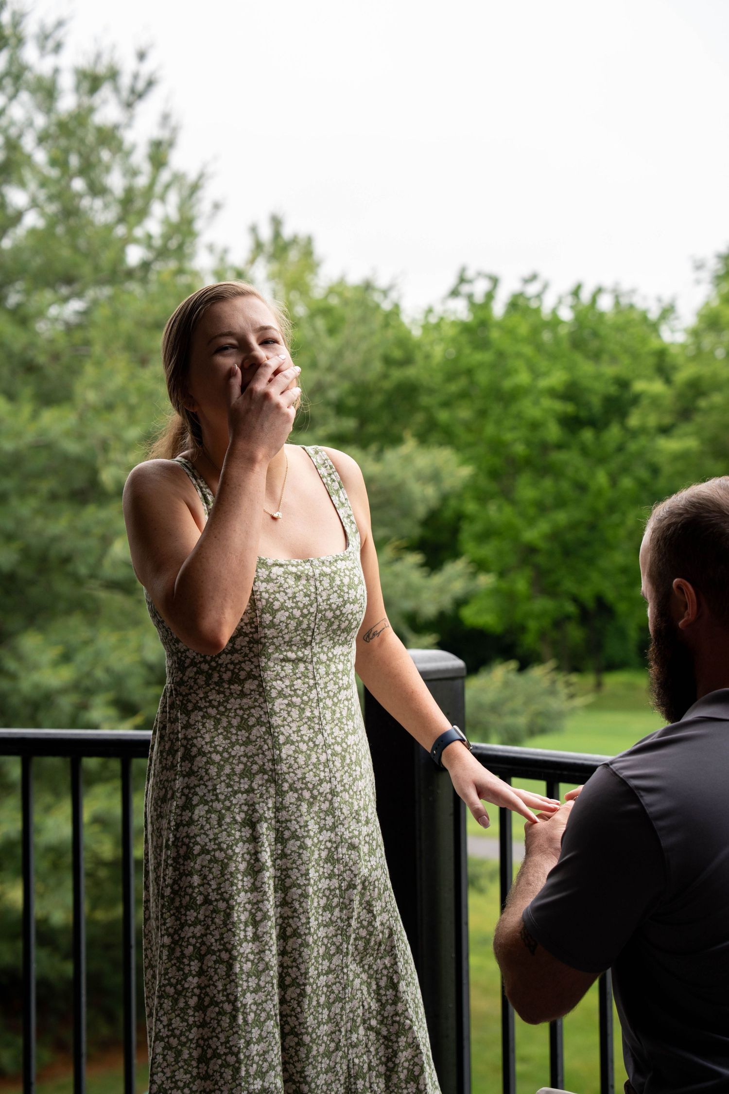 Man kneeling on outdoor deck proposing to surprised woman in floral dress with lush green trees in background.