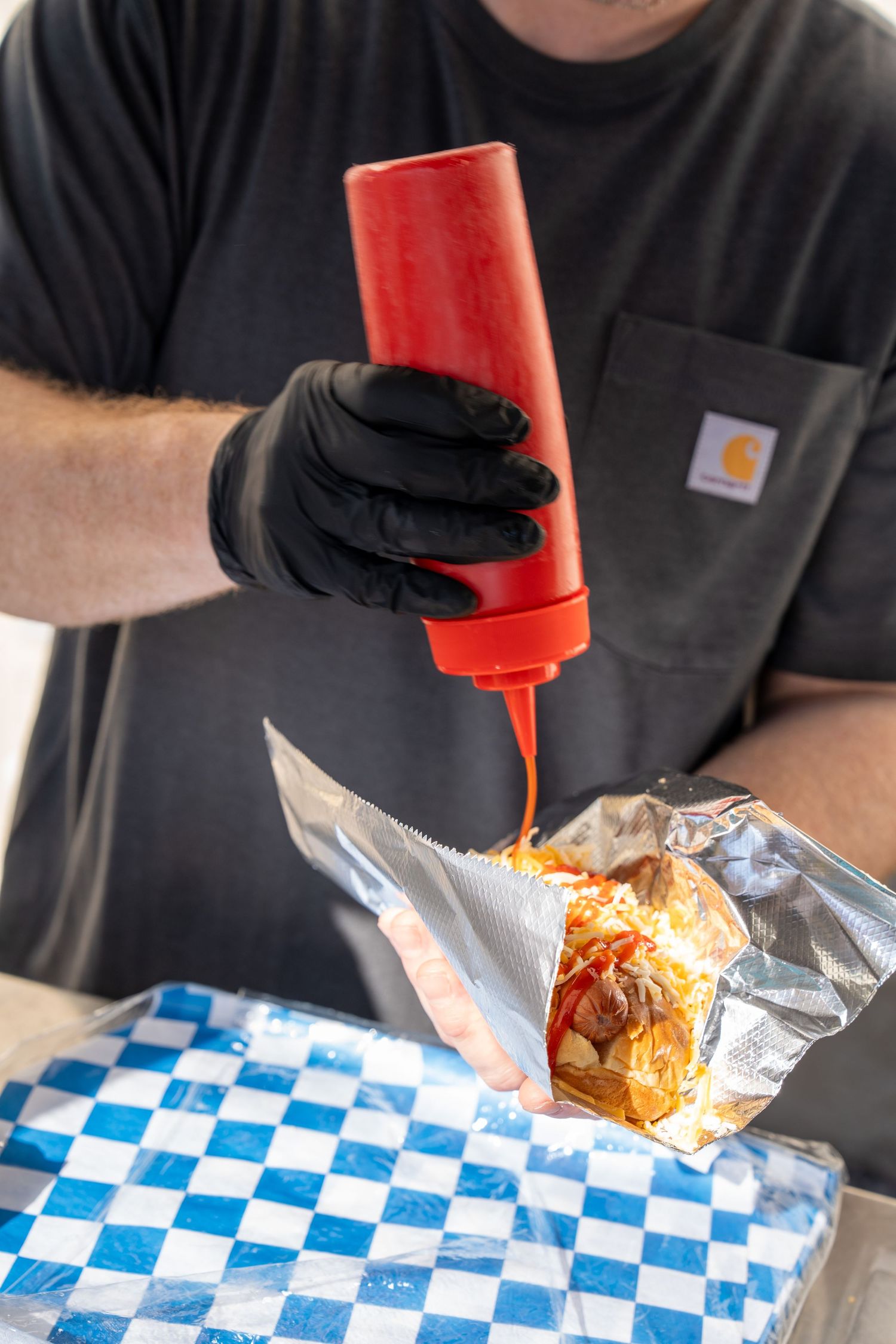 Person in black gloves squeezing red sauce onto a wrapped food item in a checkered paper-lined basket.