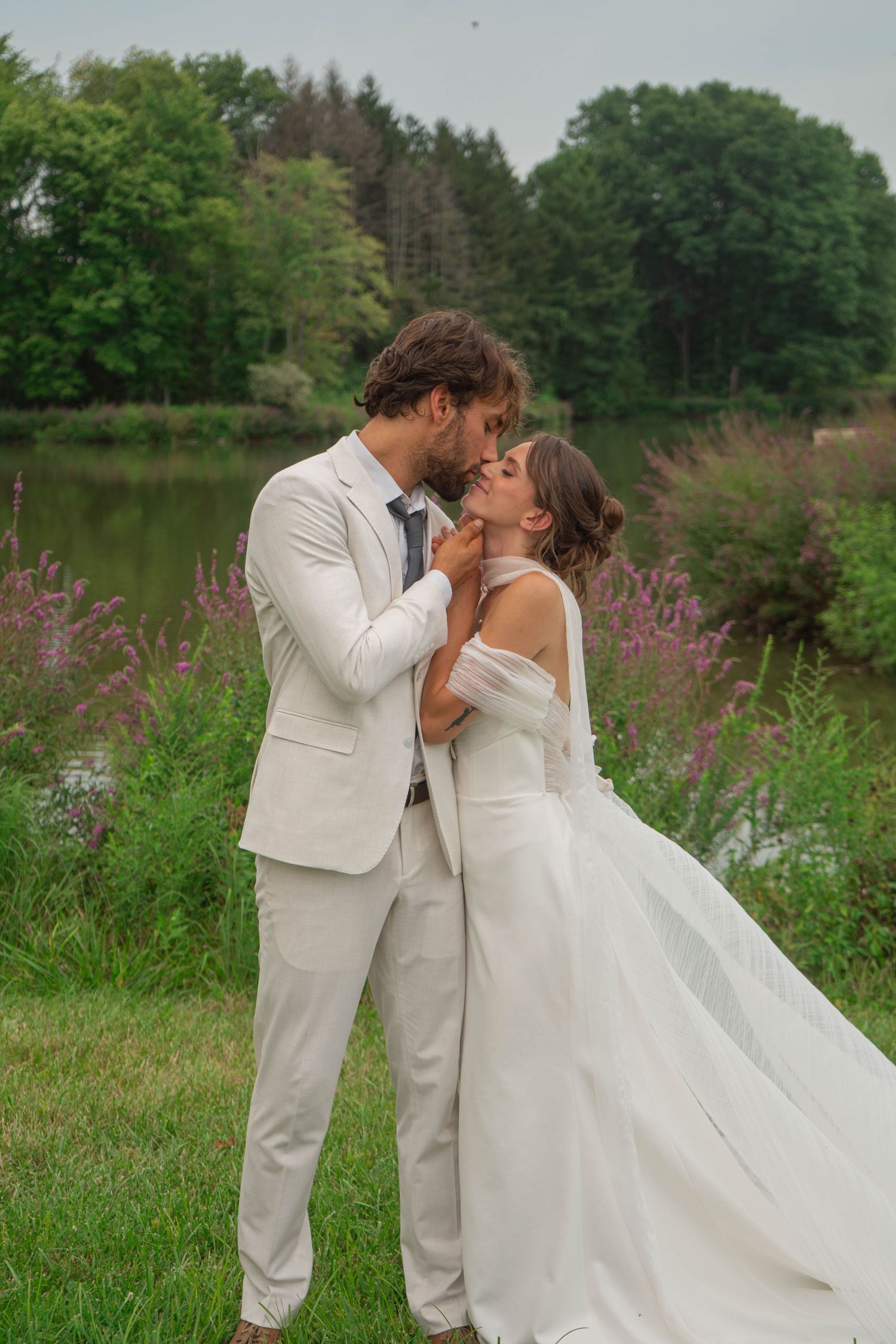 Romantic wedding couple sharing a kiss outdoors by a serene lake with lush green trees in background.