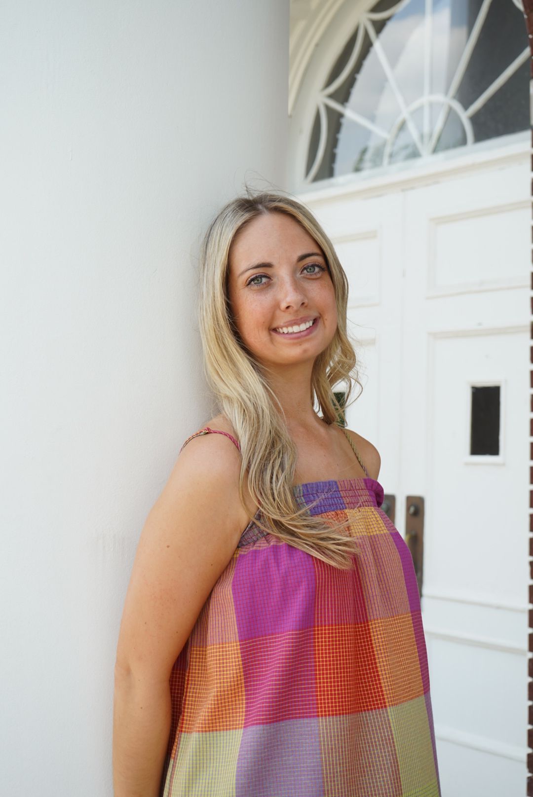 Smiling blonde woman in colorful plaid sundress leaning against white wall near door.