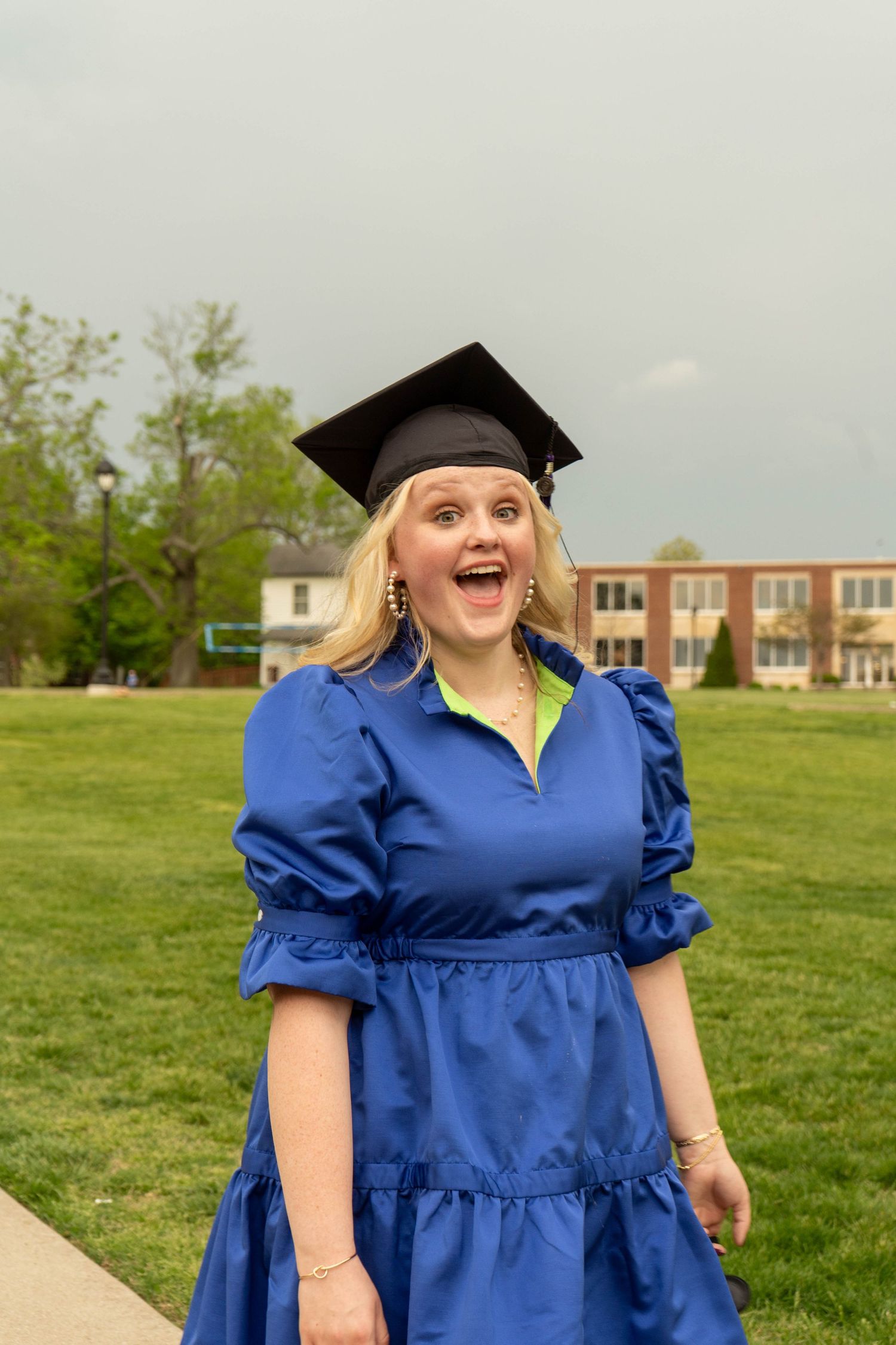 Excited blonde graduate in blue dress and mortarboard on green campus lawn.
