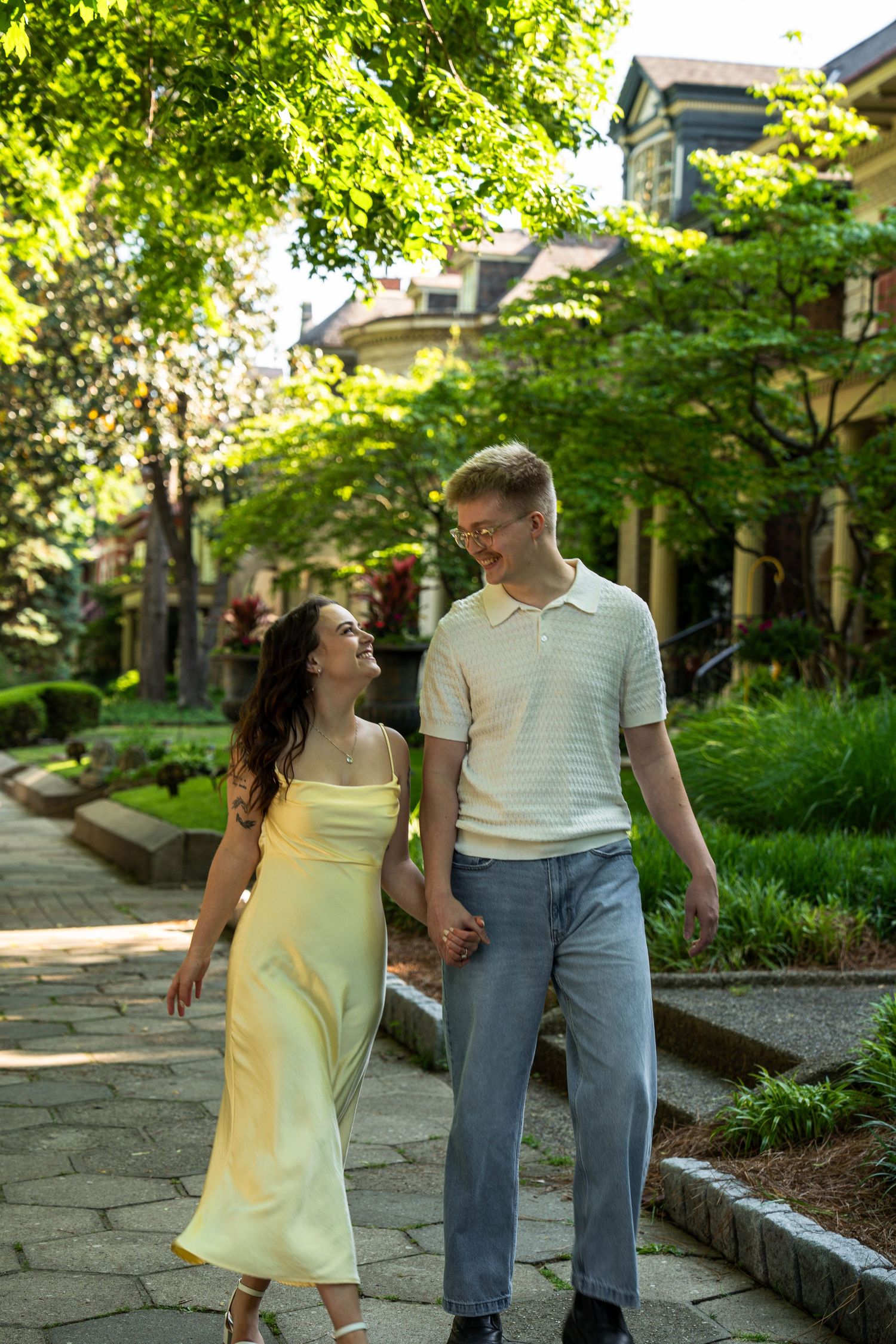Engaged couple holding hands walking along a tree-lined path in a lush neighborhood.