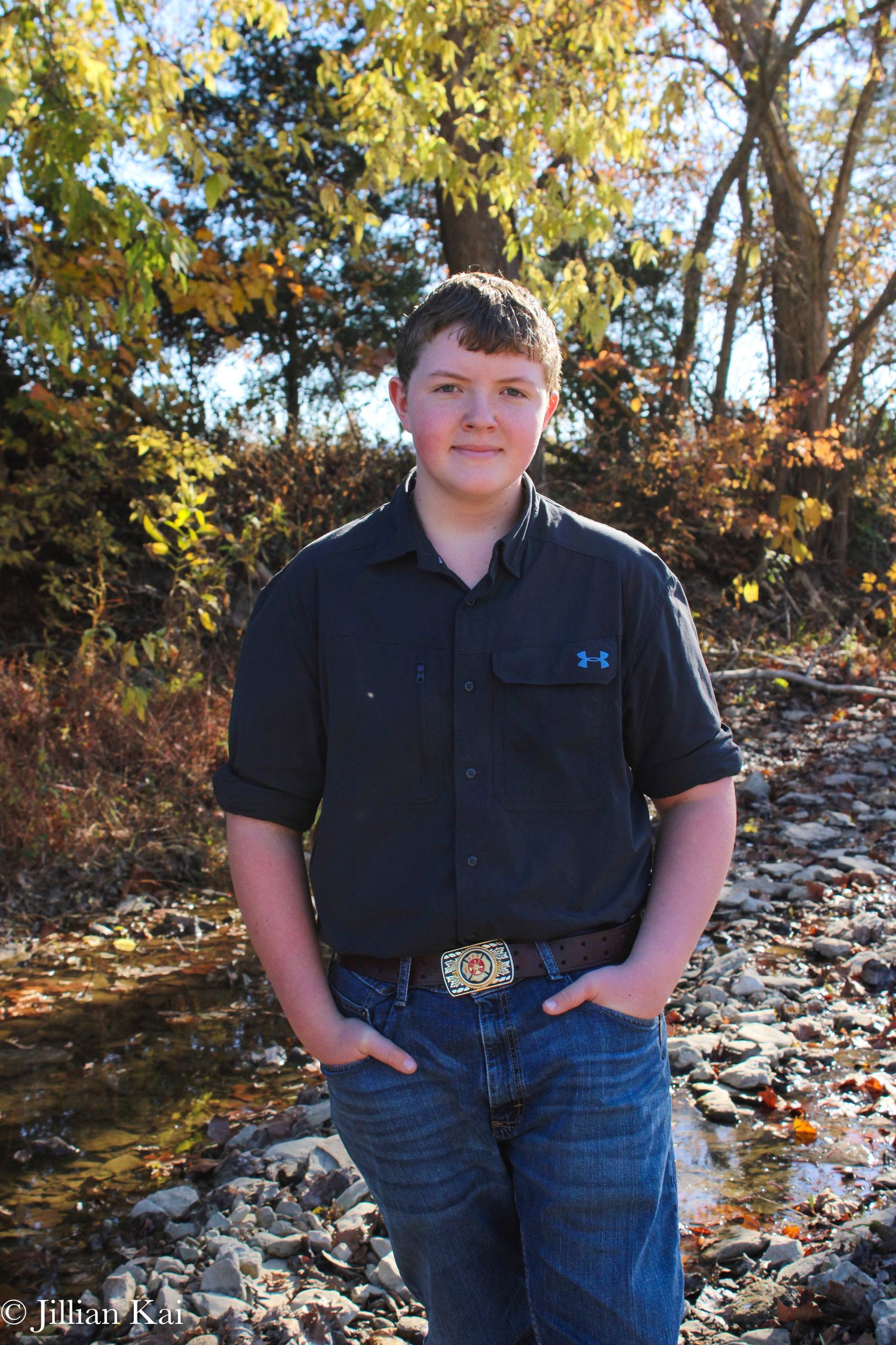 Teenage boy in dark shirt and jeans standing by creek in fall.
