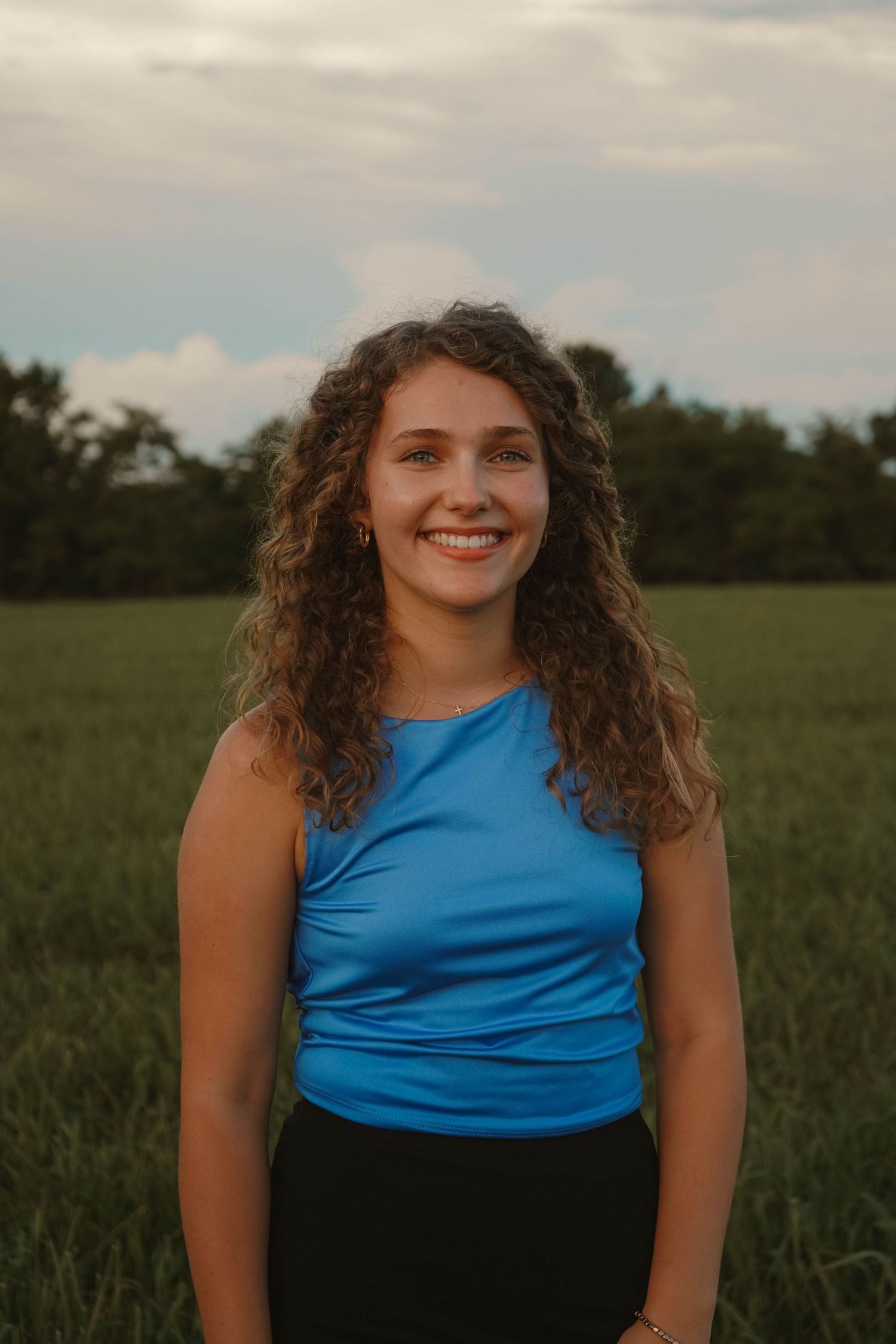Young woman with curly hair smiling in a blue sleeveless top in a green field at dusk.