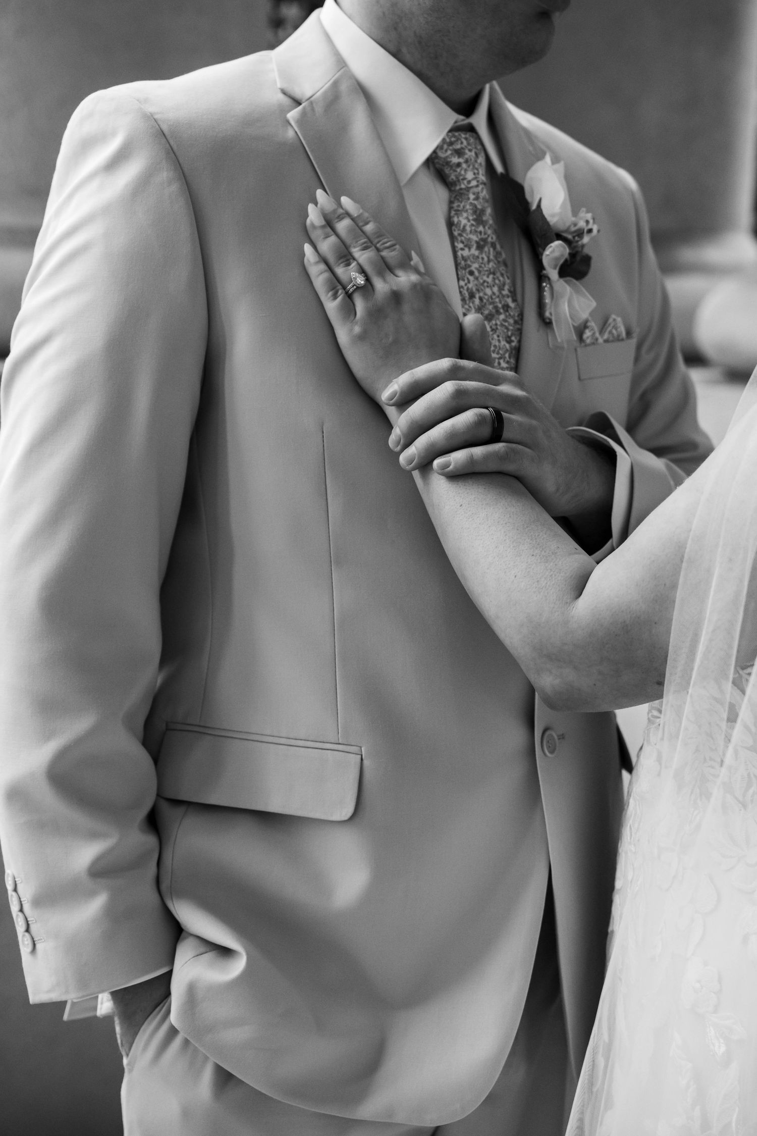 Black and white close-up of bride's hands resting on groom's tan suit jacket with boutonniere.