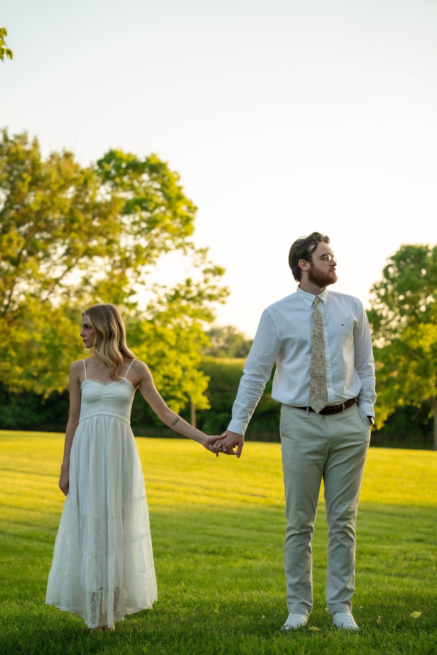 Couple holding hands in sunlit green field, woman in white dress and man in white shirt with gold tie.