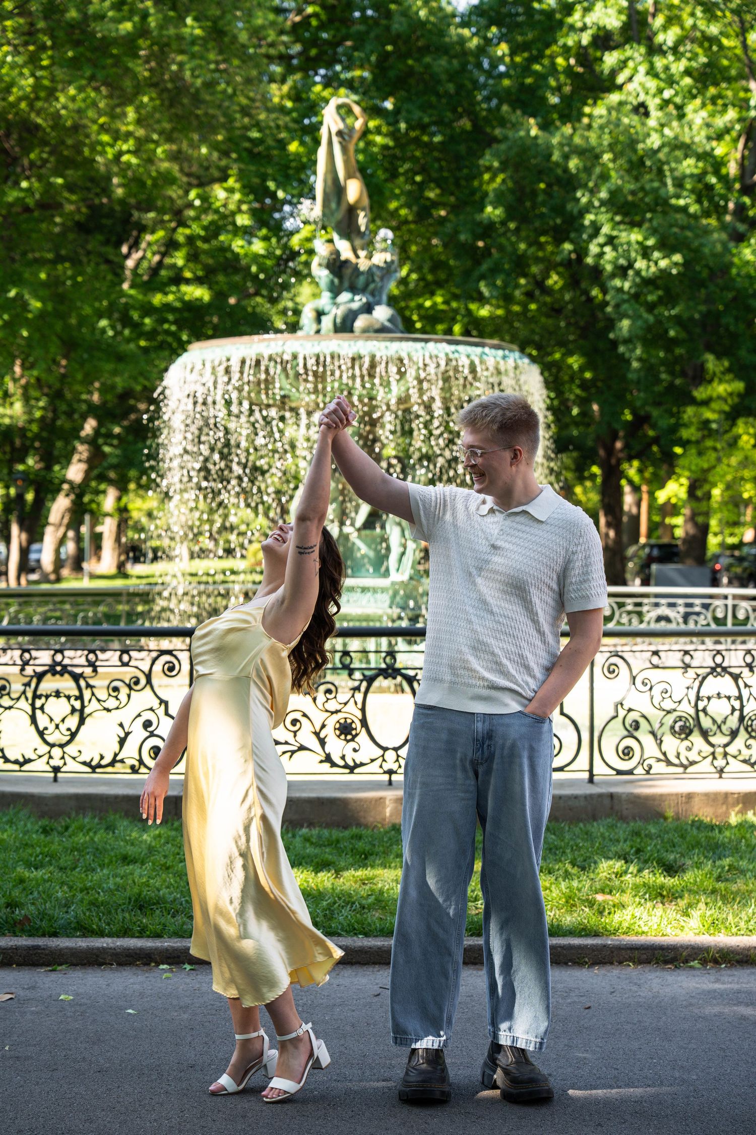Couple dancing and spinning joyfully in front of a decorative fountain in a sunny park.