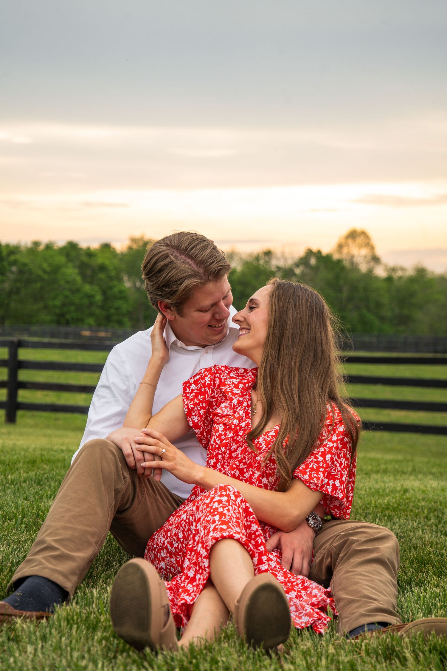 Couple sitting on grass at sunset, woman in red floral dress, man in white shirt, embracing romantically.