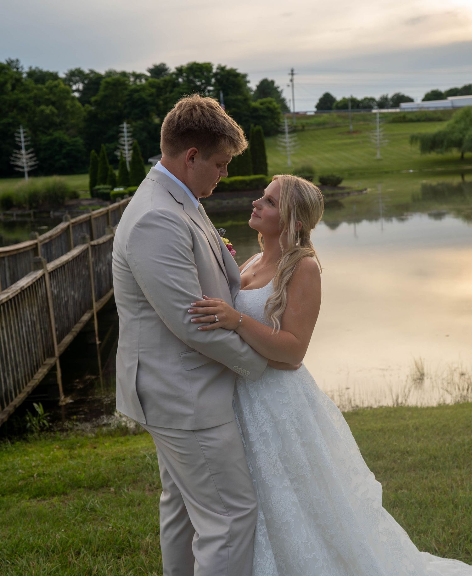 Bride and groom share a romantic moment by a pond at sunset, dressed in a white gown and light gray suit.