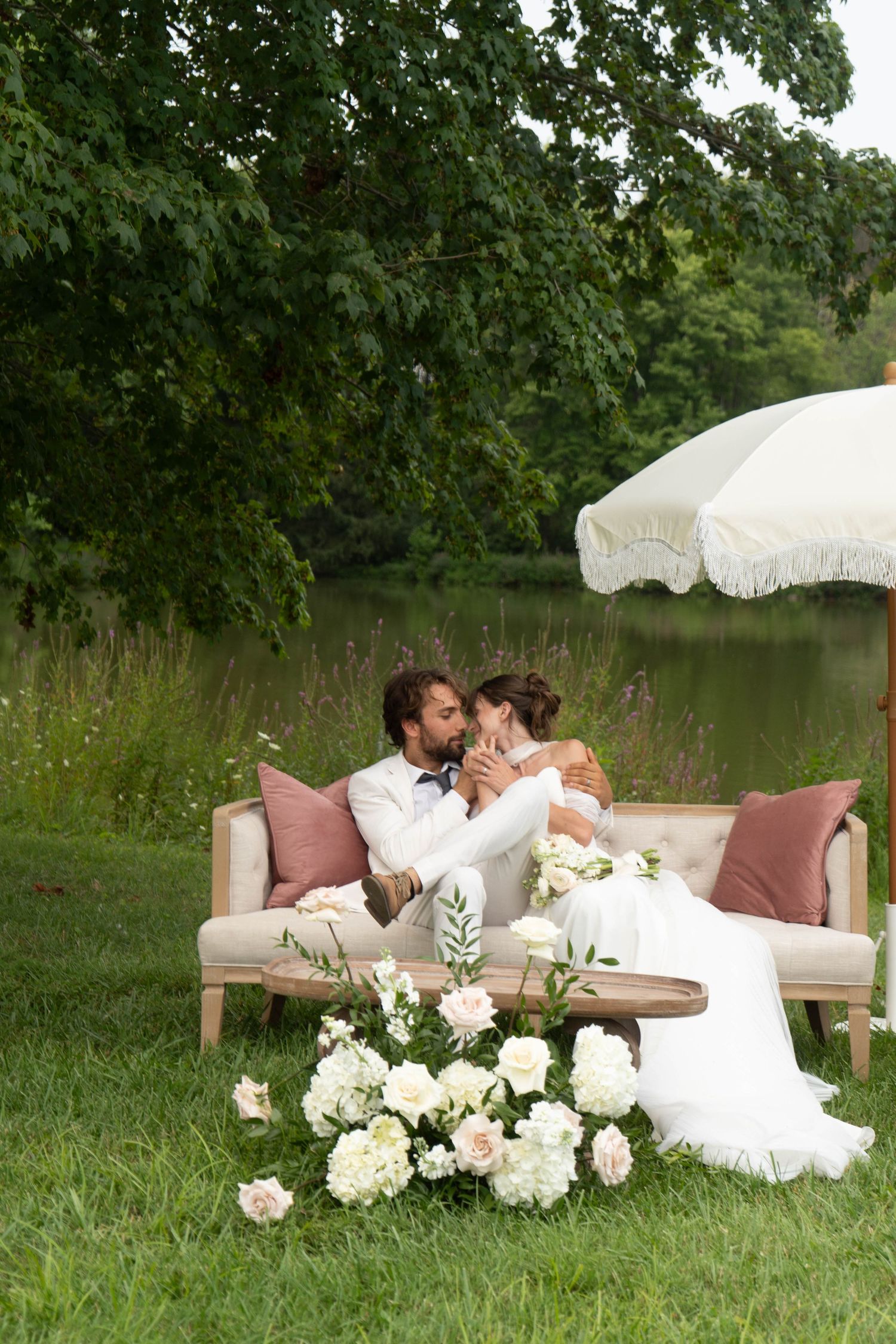 Bride and groom share a kiss on an outdoor sofa surrounded by white roses and a fringed umbrella.