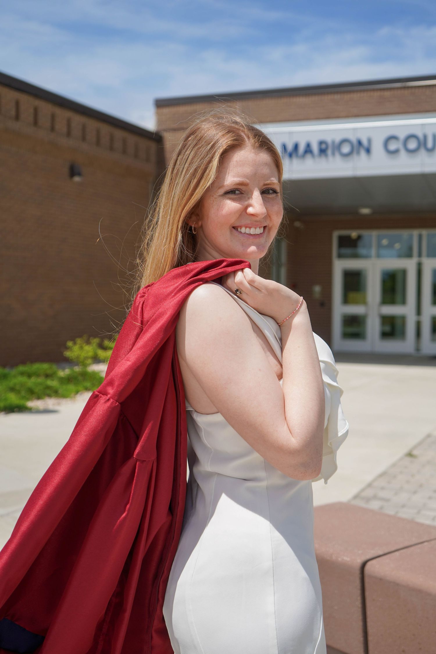 Smiling young woman in white dress carries red graduation gown over shoulder outside Marion County High School.
