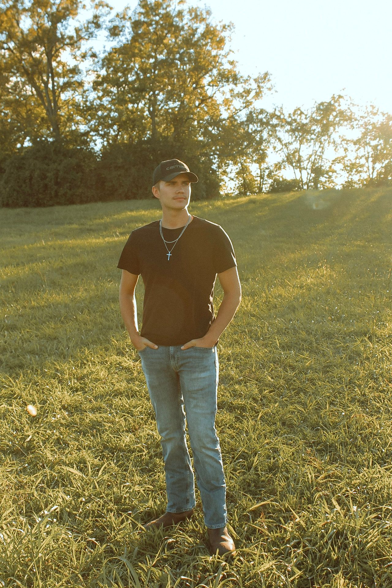 Young man in black t-shirt and cap stands in golden sunlit grassy field with trees.