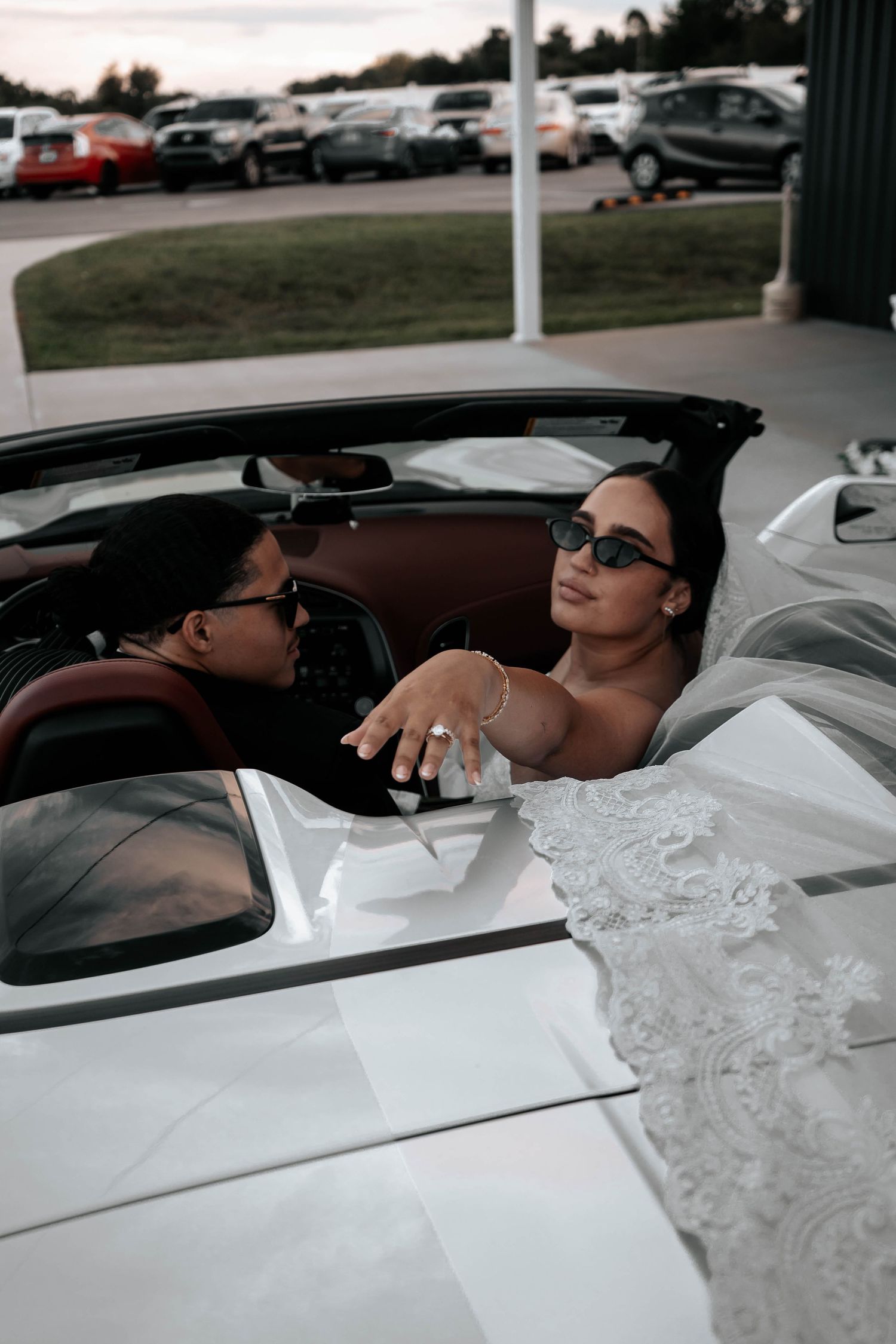 Bride and groom wearing sunglasses share a fun moment in white convertible sports car after wedding.