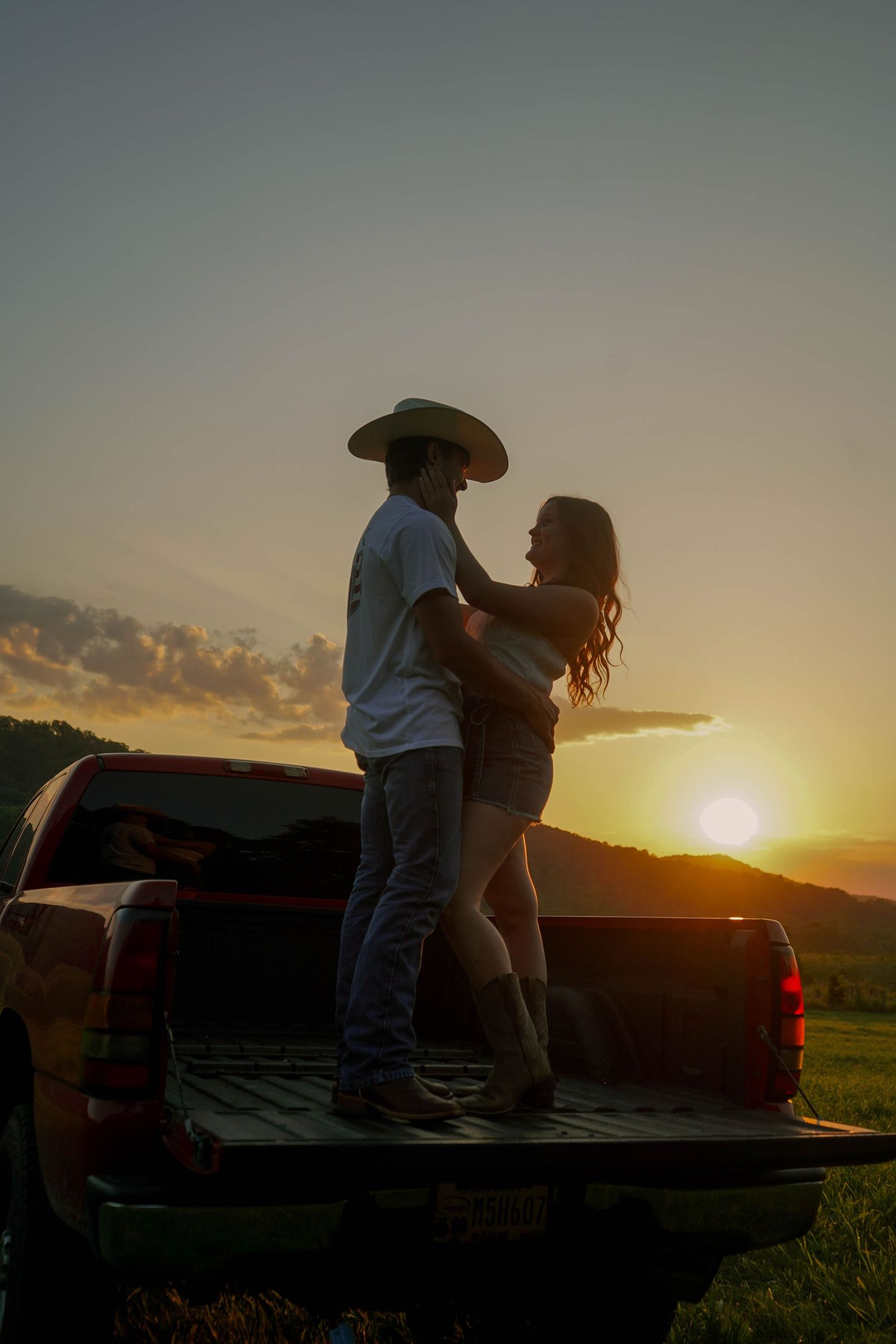 Silhouette of couple standing and embracing on a truck bed at sunset with glowing orange sky over green hills.