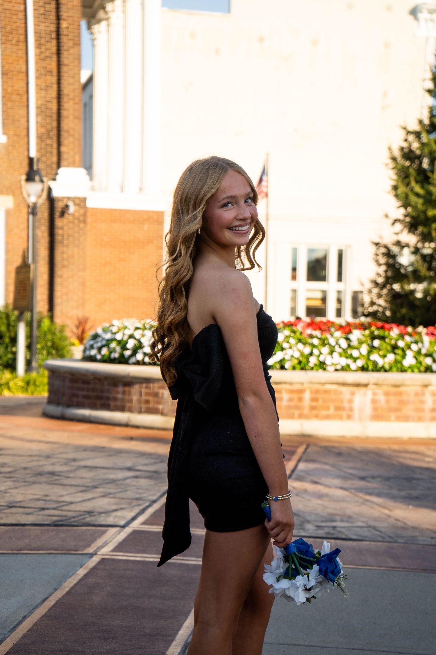 Smiling blonde teen in strapless black mini dress holds blue and white flowers outdoors.