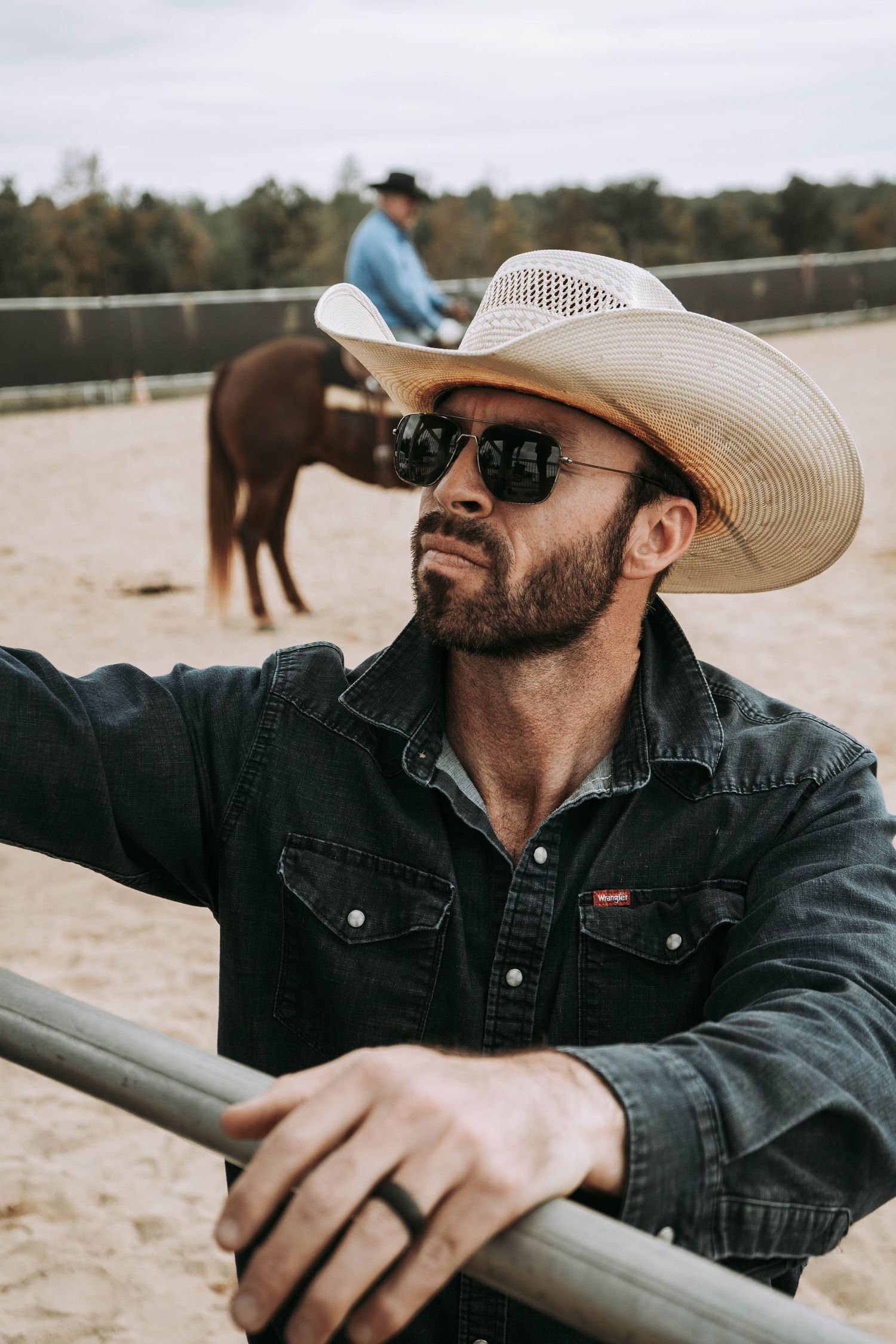 Cowboy leaning on metal fence rail in white hat and sunglasses with horse and rider behind him.