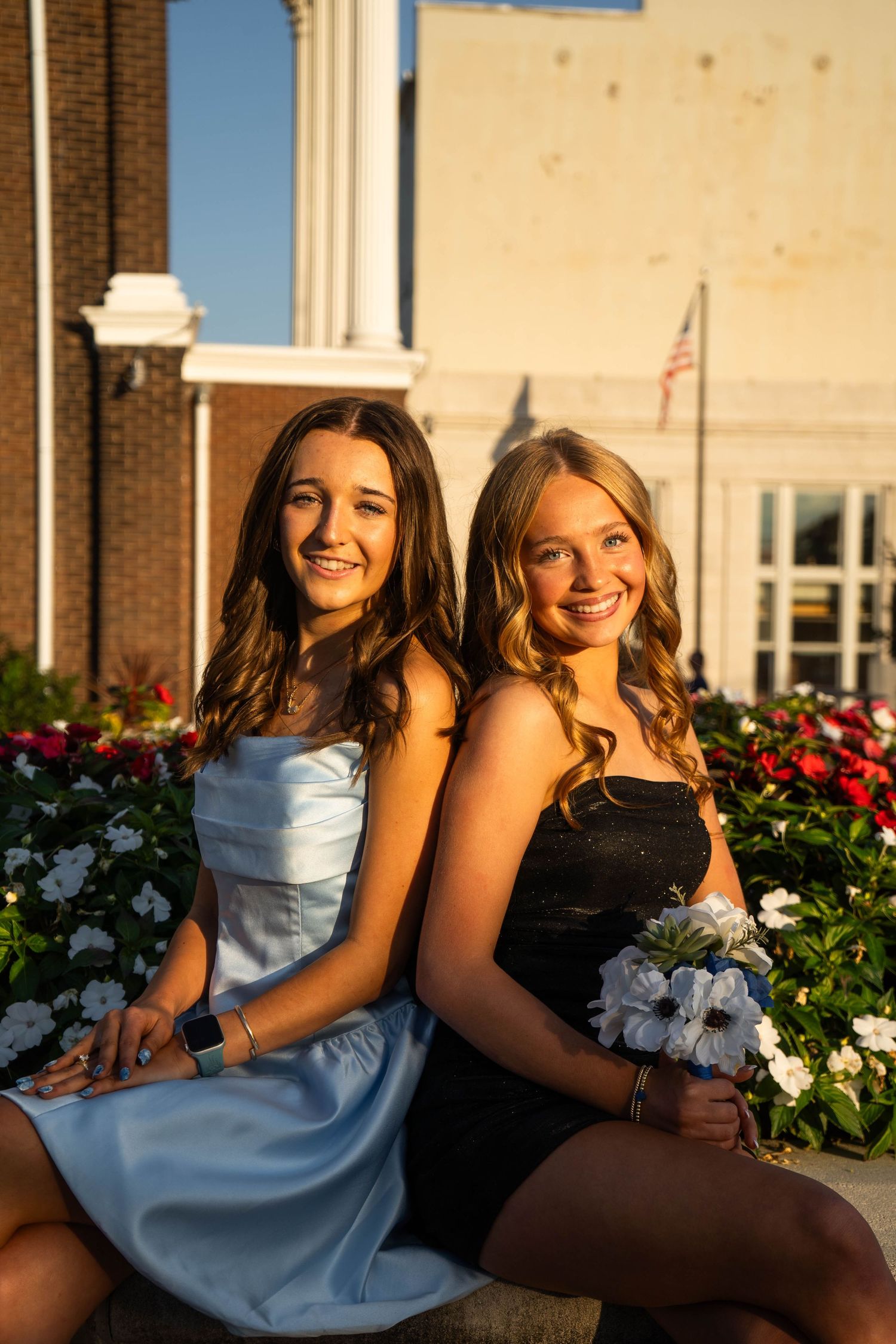 Two teenage girls in formal dresses sit back-to-back near a flower garden at golden hour.