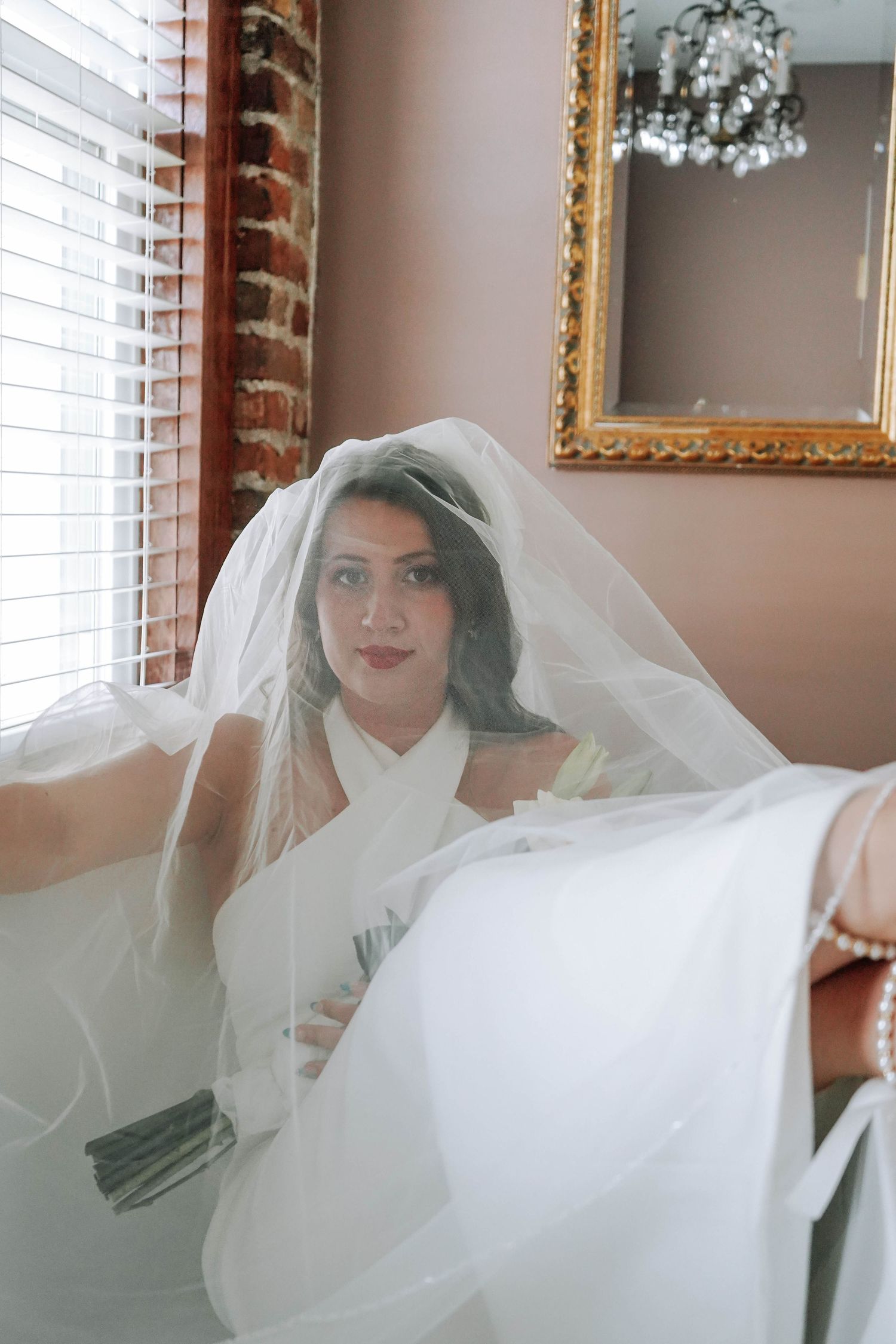 Bride in white wedding dress and flowing veil spreads arms wide in elegant room with gold-framed mirror.