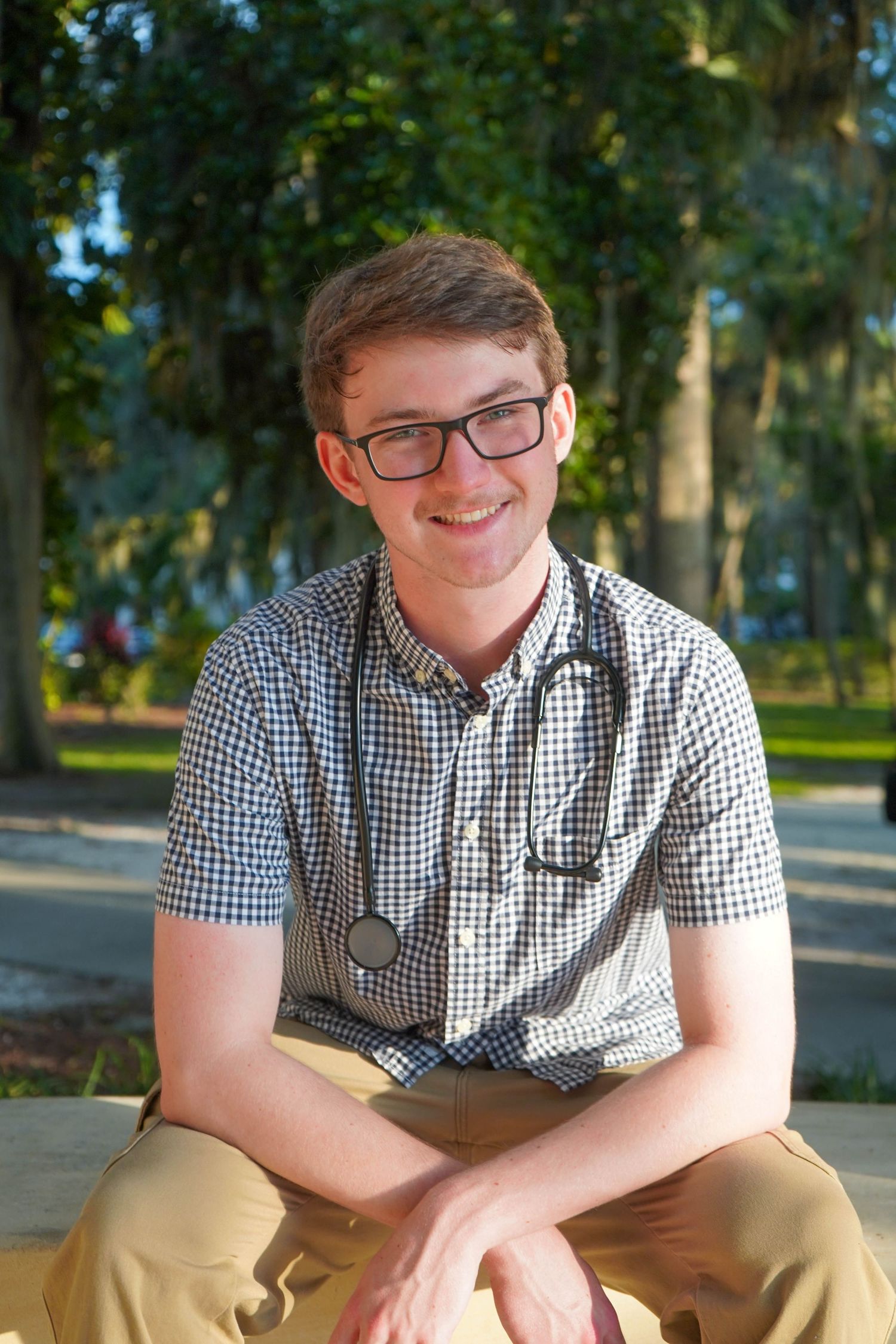 Smiling young medical student with stethoscope and glasses sitting outdoors in a sunny park setting.