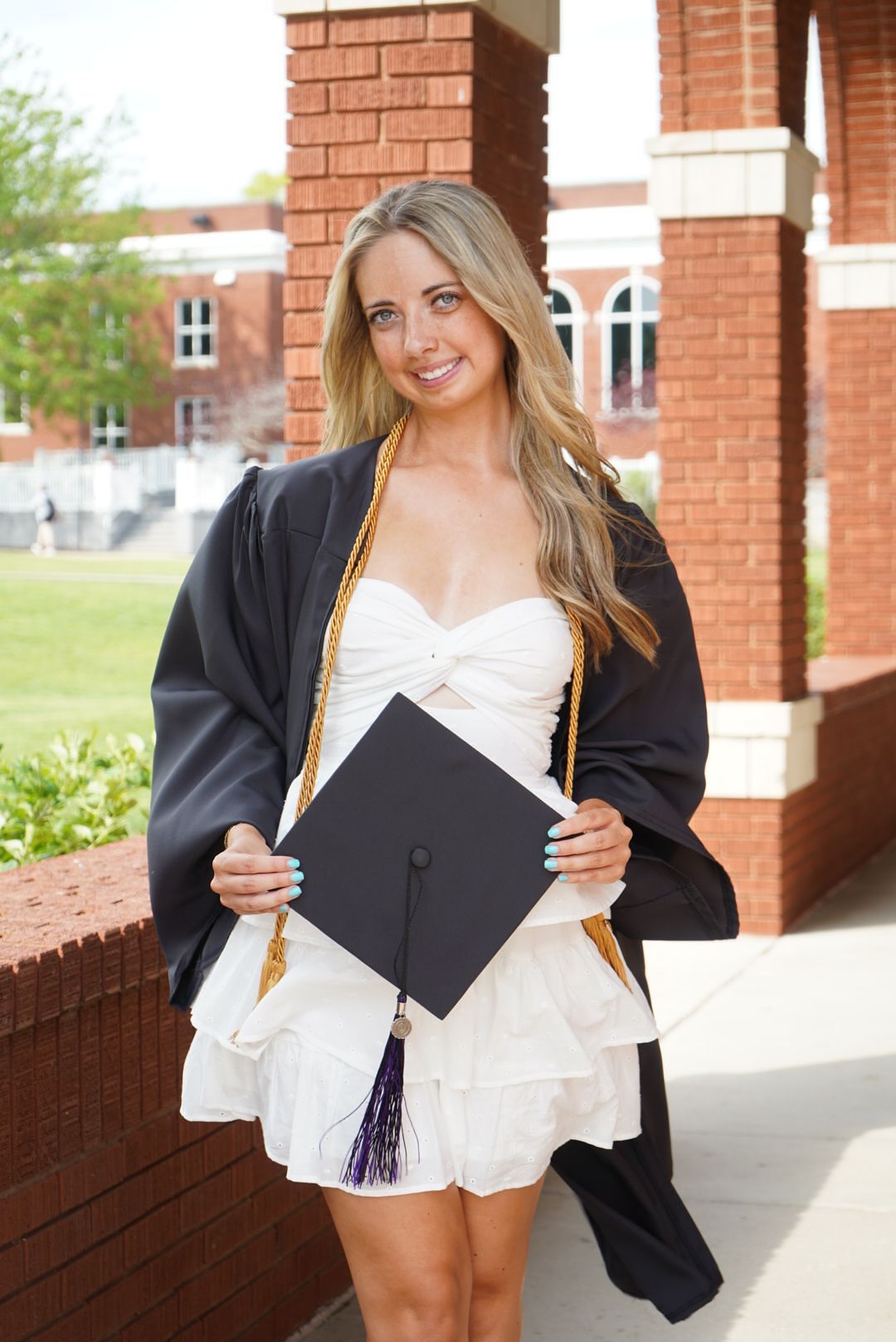 Smiling blonde graduate in white dress holding cap and black gown near brick building.