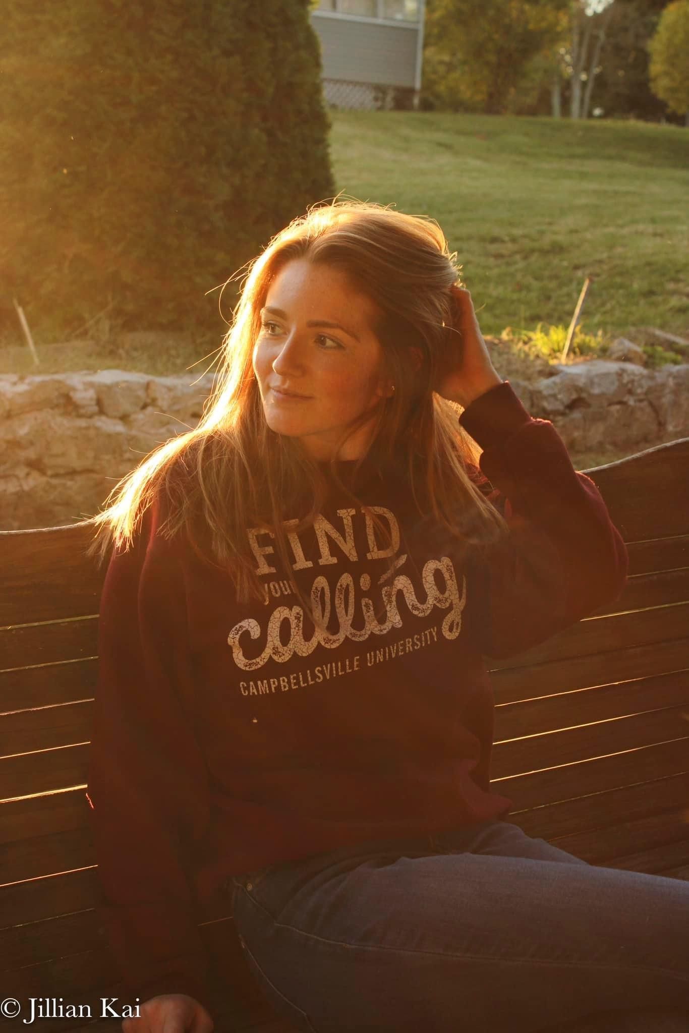 Woman in Find Your Calling Campbellsville University hoodie sitting on bench in warm golden sunlight.