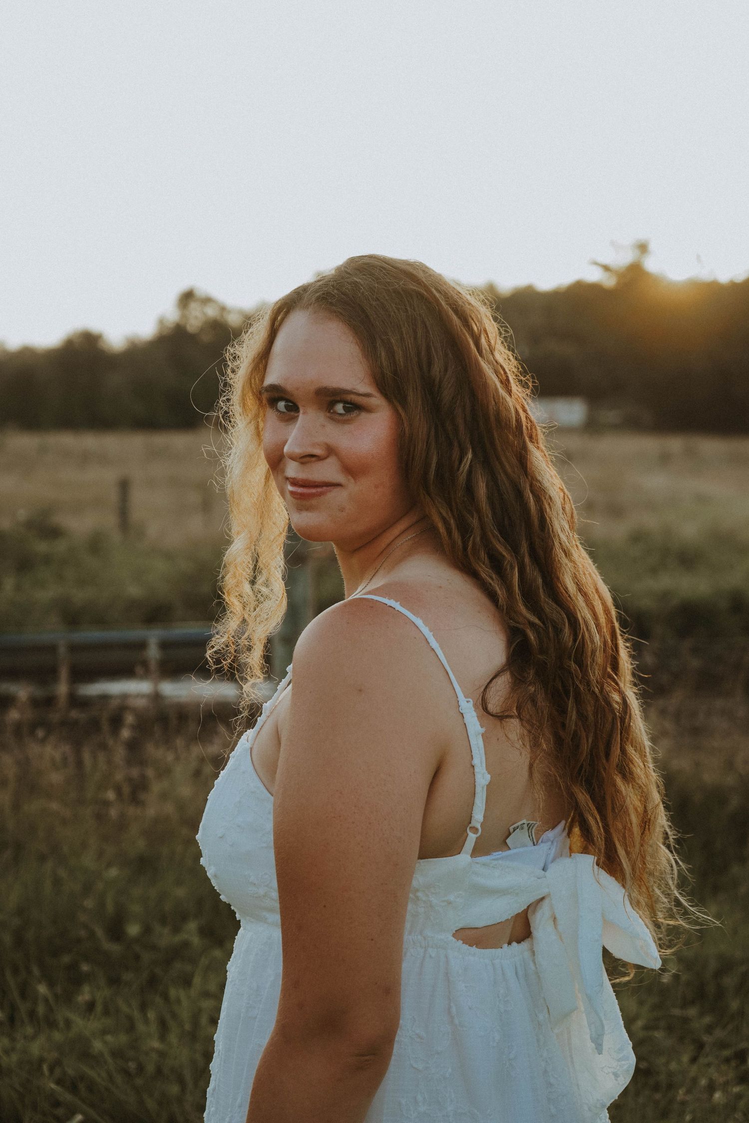 Young woman in white sundress smiling over shoulder at golden hour in a farm field.