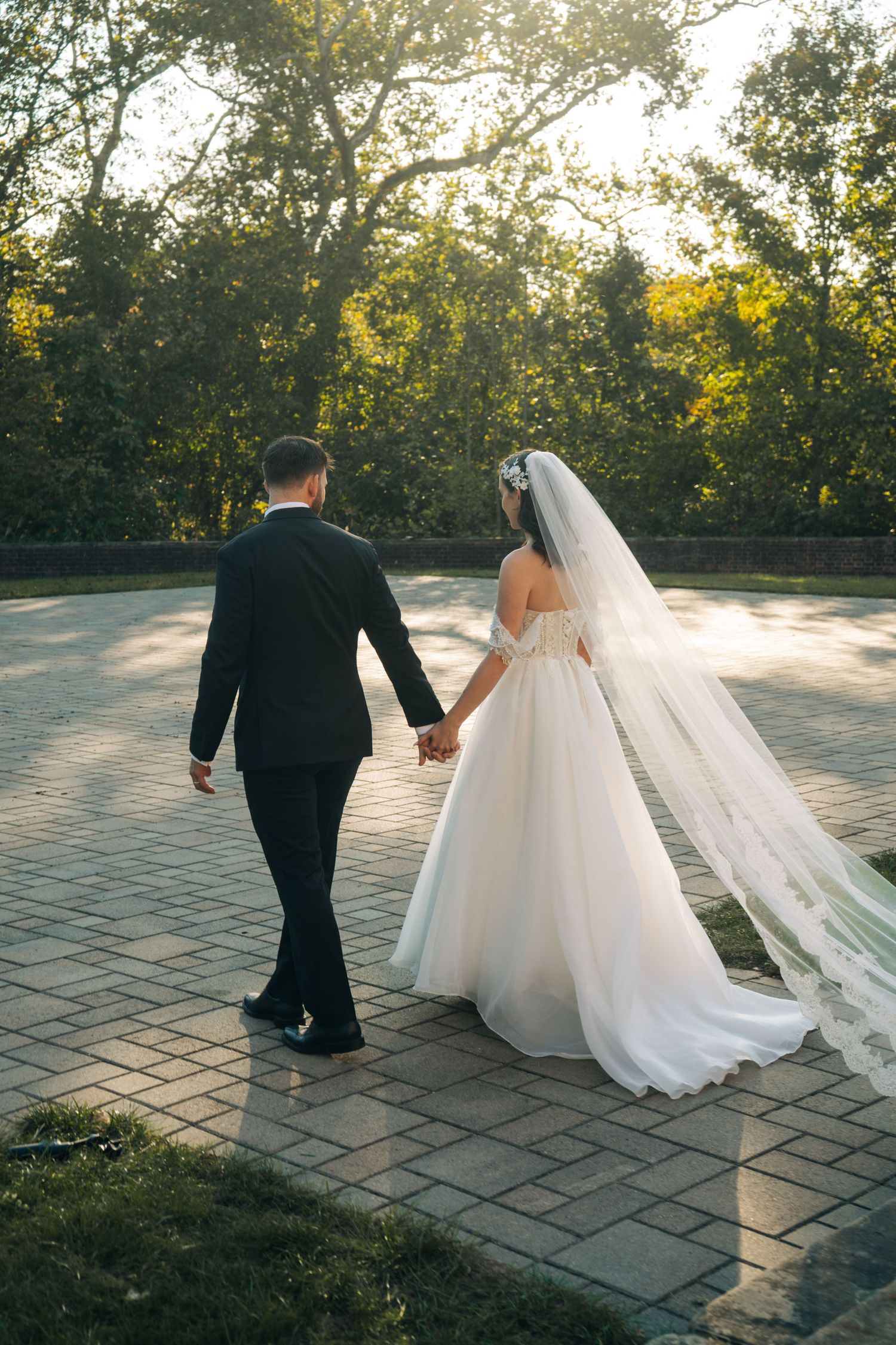 Bride and groom walking hand in hand on a brick path surrounded by lush green trees during golden hour.