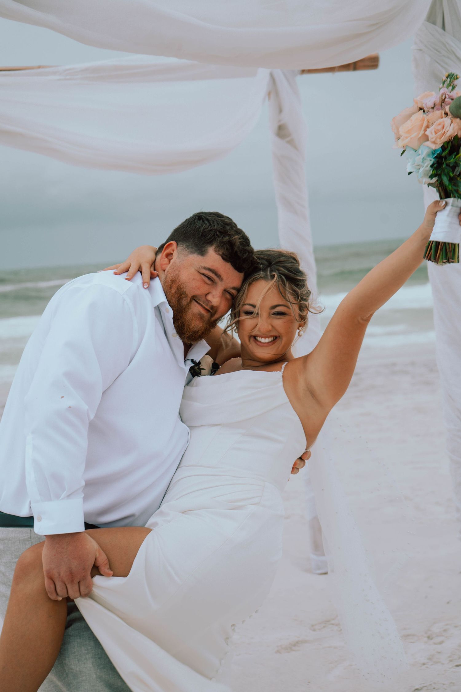 Joyful bride and groom celebrating on the beach under a draped canopy with a pink bouquet.
