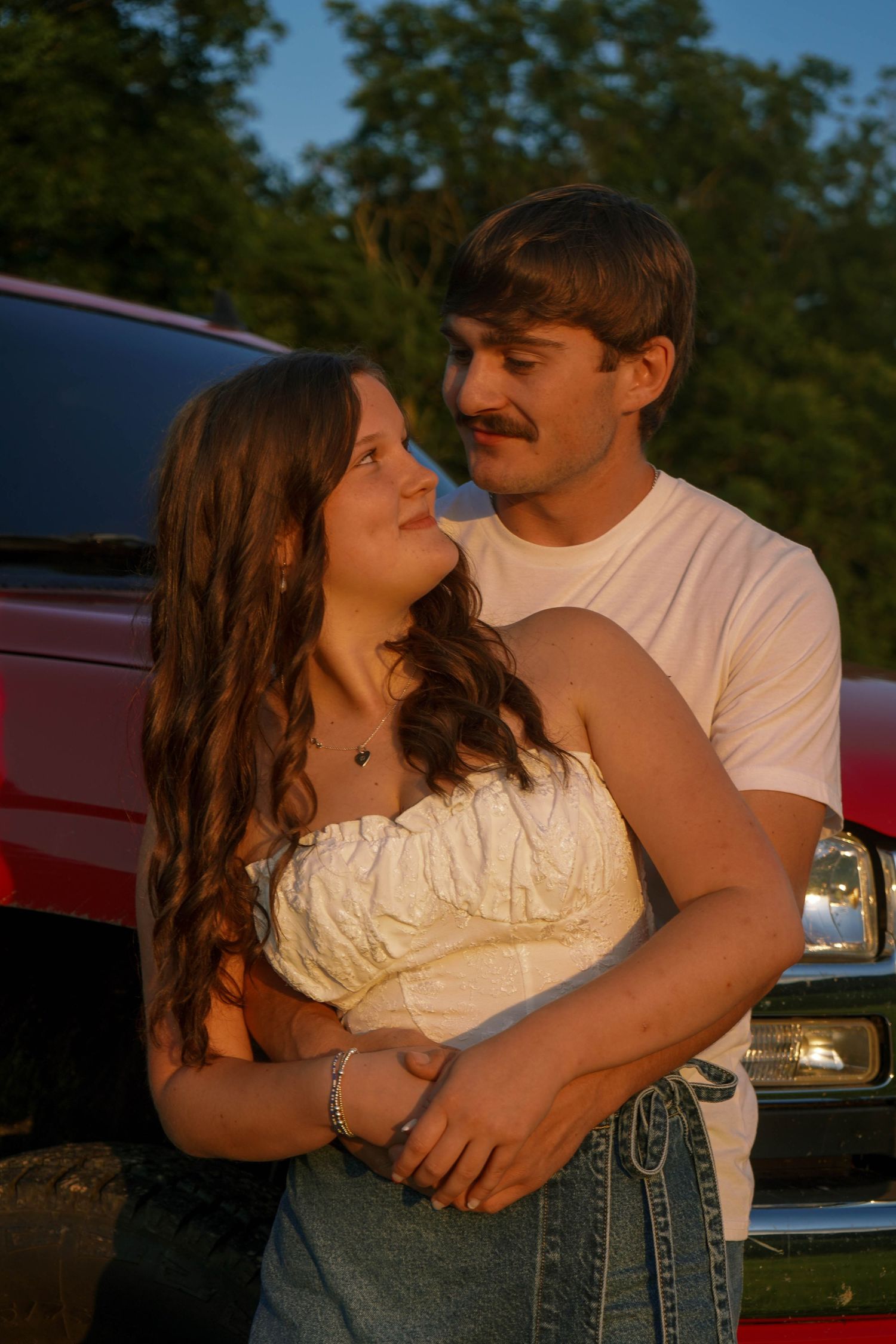 Young couple in white shirts embracing romantically in front of a red truck at golden hour sunset.