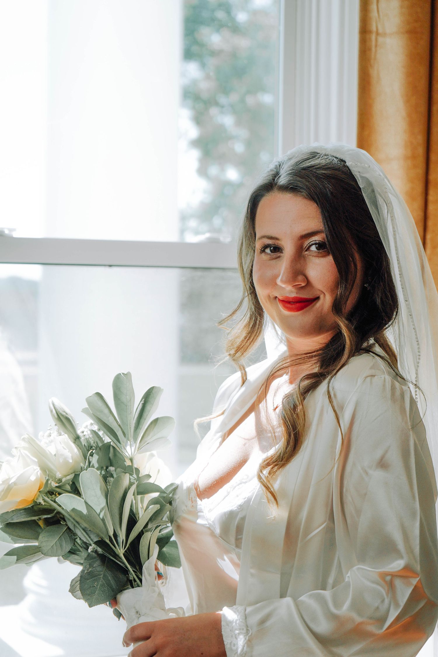 Radiant bride with red lips in white robe and veil smiles holding white flower bouquet by bright window.