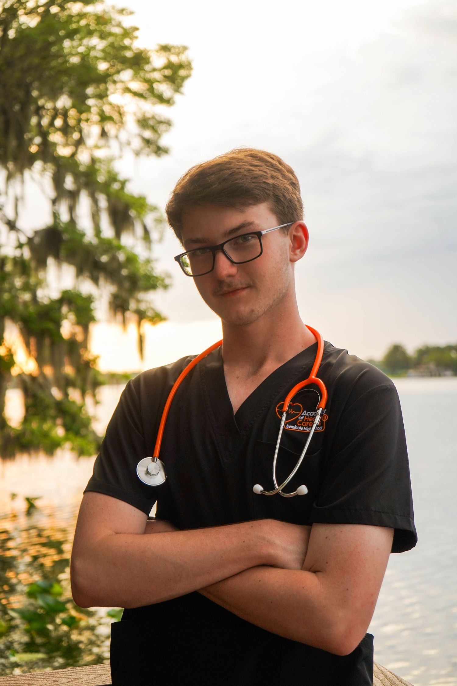 Confident young medical professional in black scrubs with orange stethoscope posing by a lake at sunset.