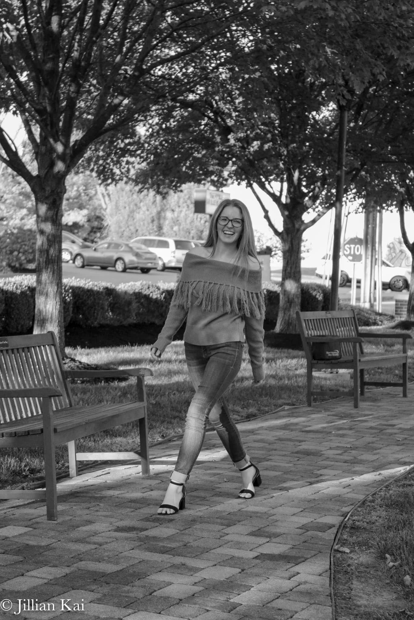 Smiling woman in fringe sweater and glasses walking on brick path between benches in black and white.