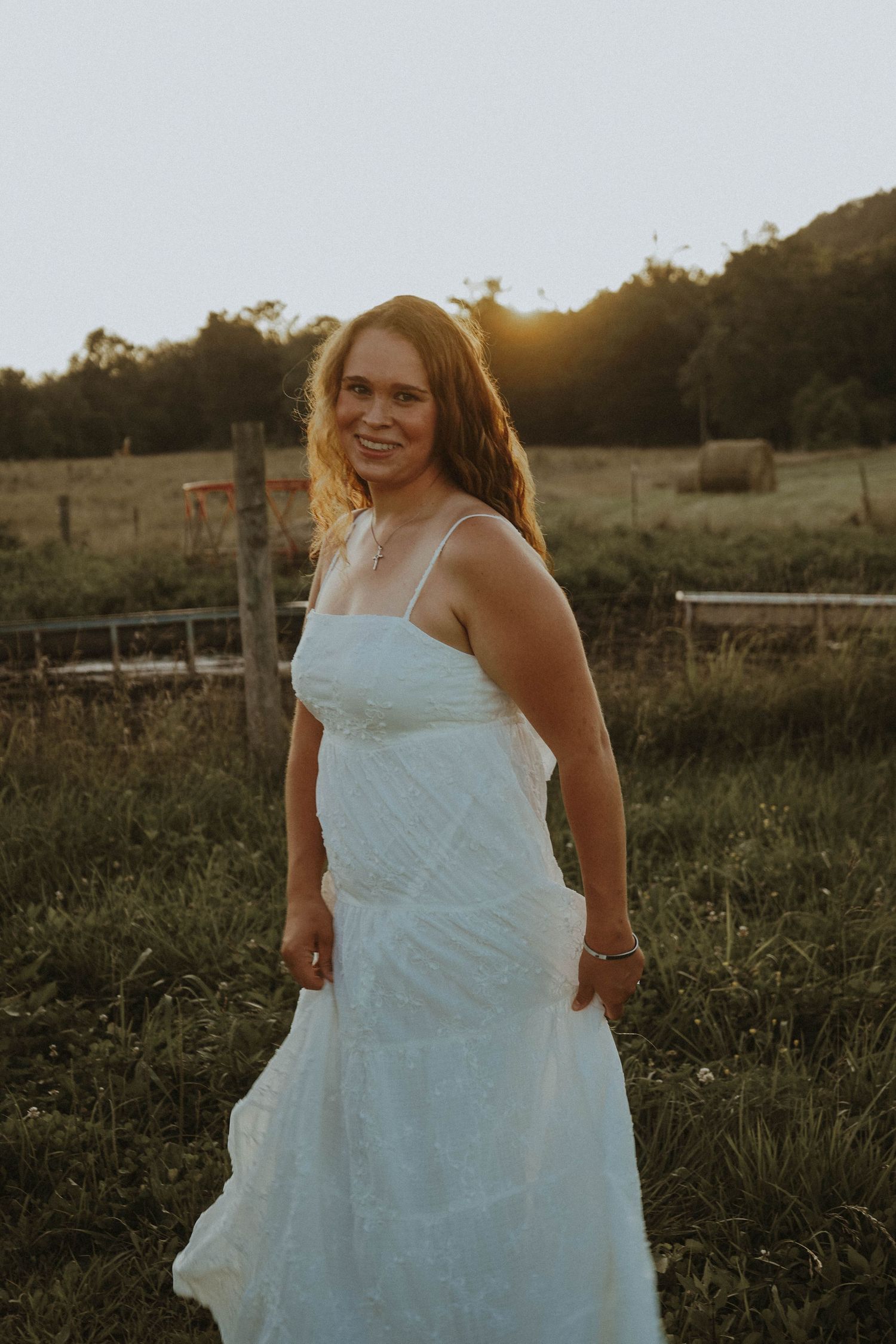 Young woman in white lace dress twirling in a green farm field at sunset.