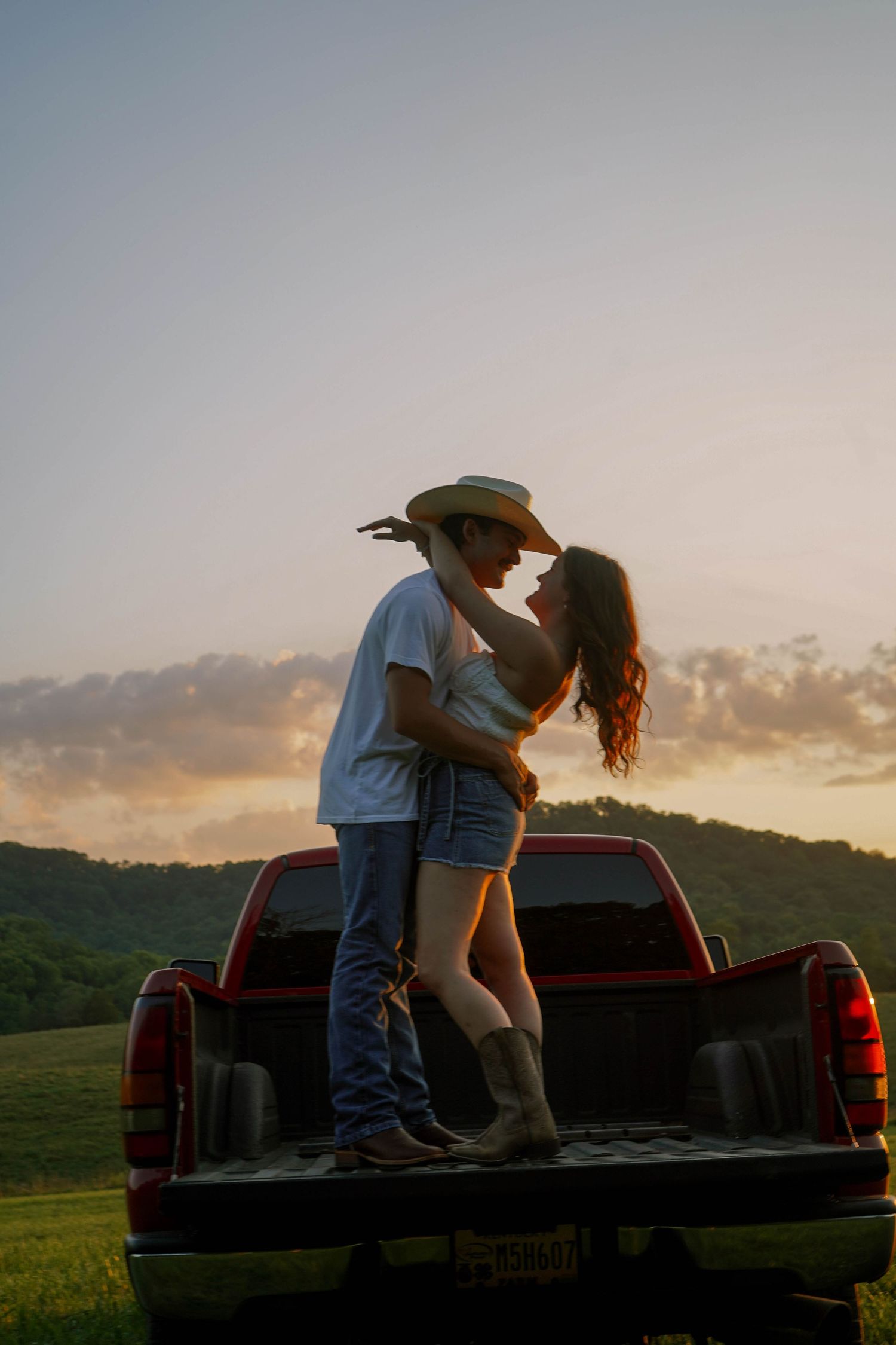 Romantic couple in cowboy hats embracing on a truck bed at dusk with scenic rolling hills and sunset sky behind them.