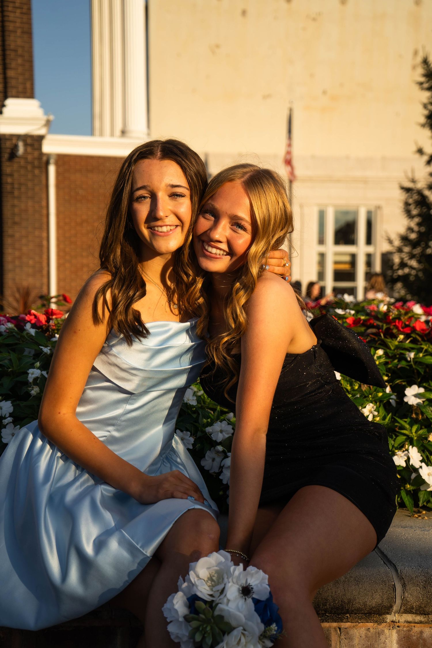 Two smiling teenage girls in formal dresses pose together by colorful flowers at sunset.
