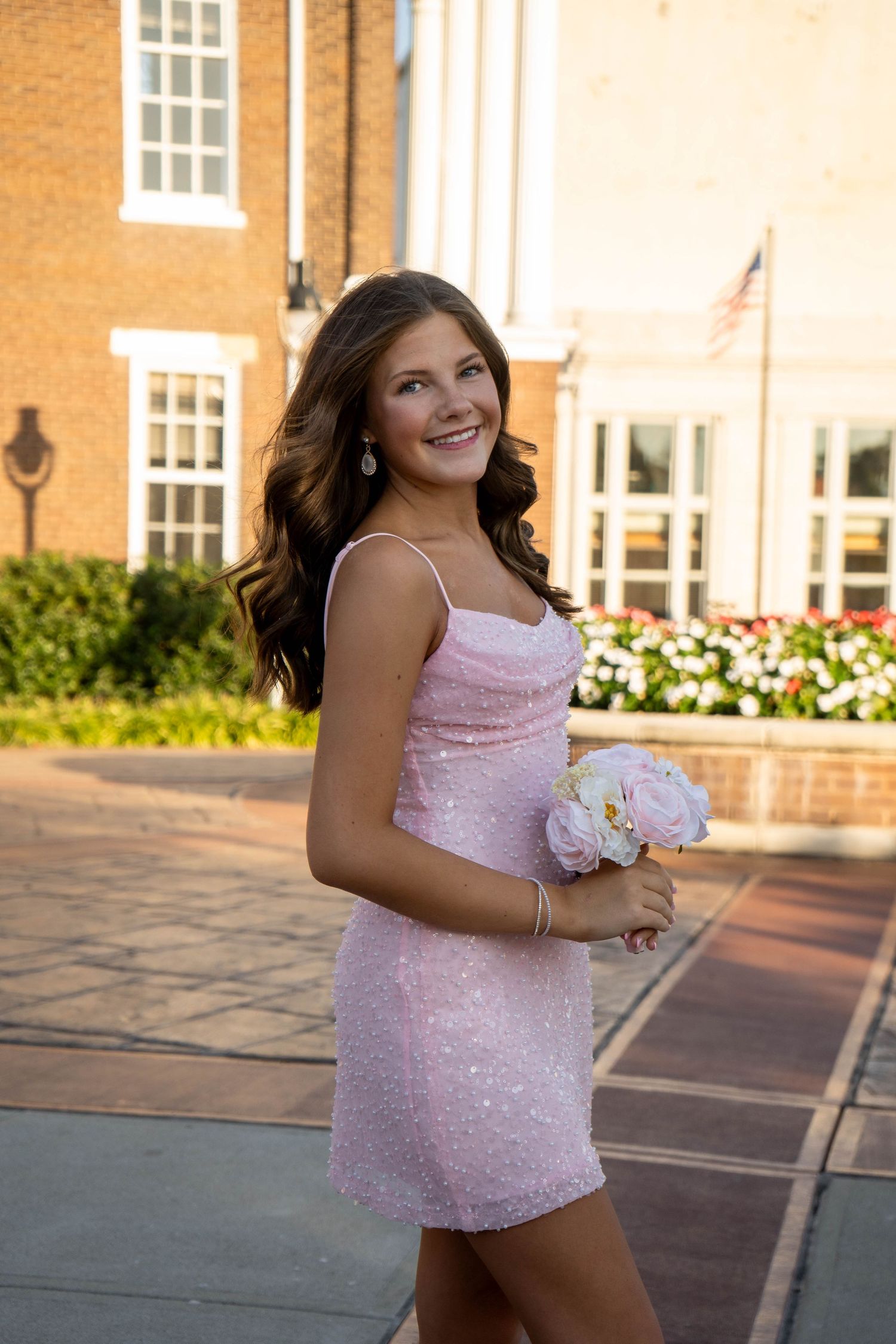 Smiling brunette teen in pink sequin dress holds white flowers outside brick building at sunset.