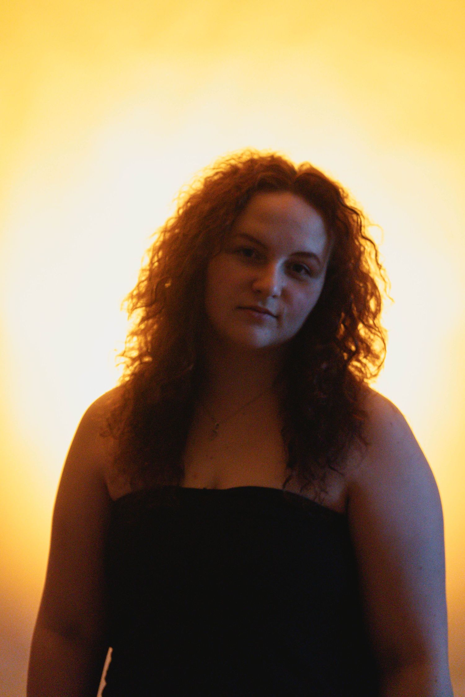 Young woman with curly brown hair wearing black top posing against warm golden yellow background.