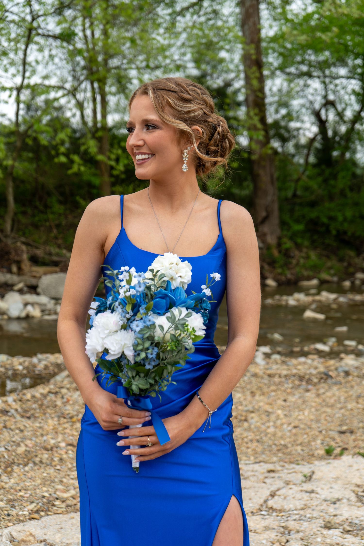 Elegant young woman in royal blue prom dress holding white and blue floral bouquet outdoors by a creek.