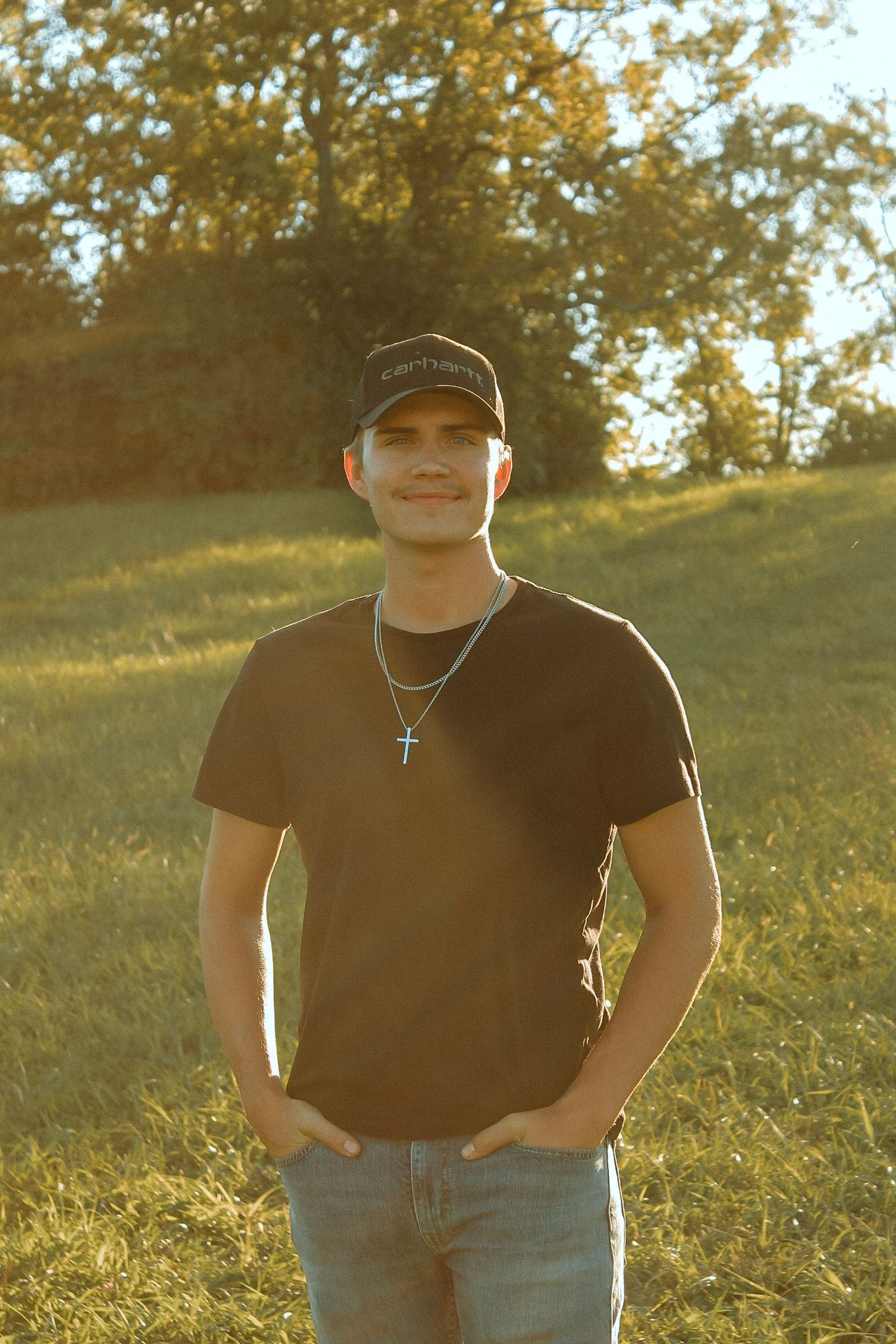 Smiling young man wearing black Carhartt cap and cross necklace poses in sunny green field.