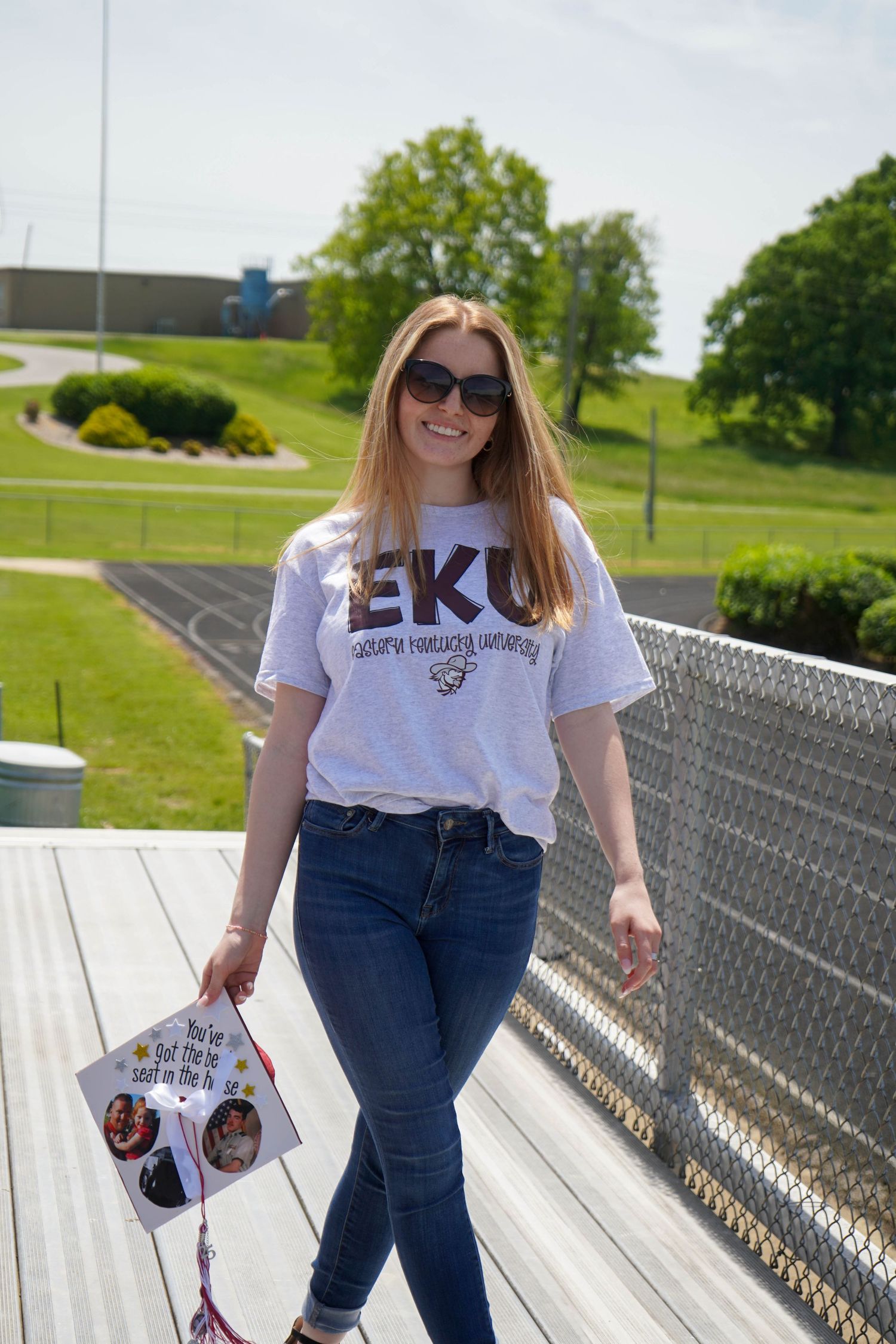 Smiling young woman in EKU Eastern Kentucky University shirt holds graduation cap outdoors.